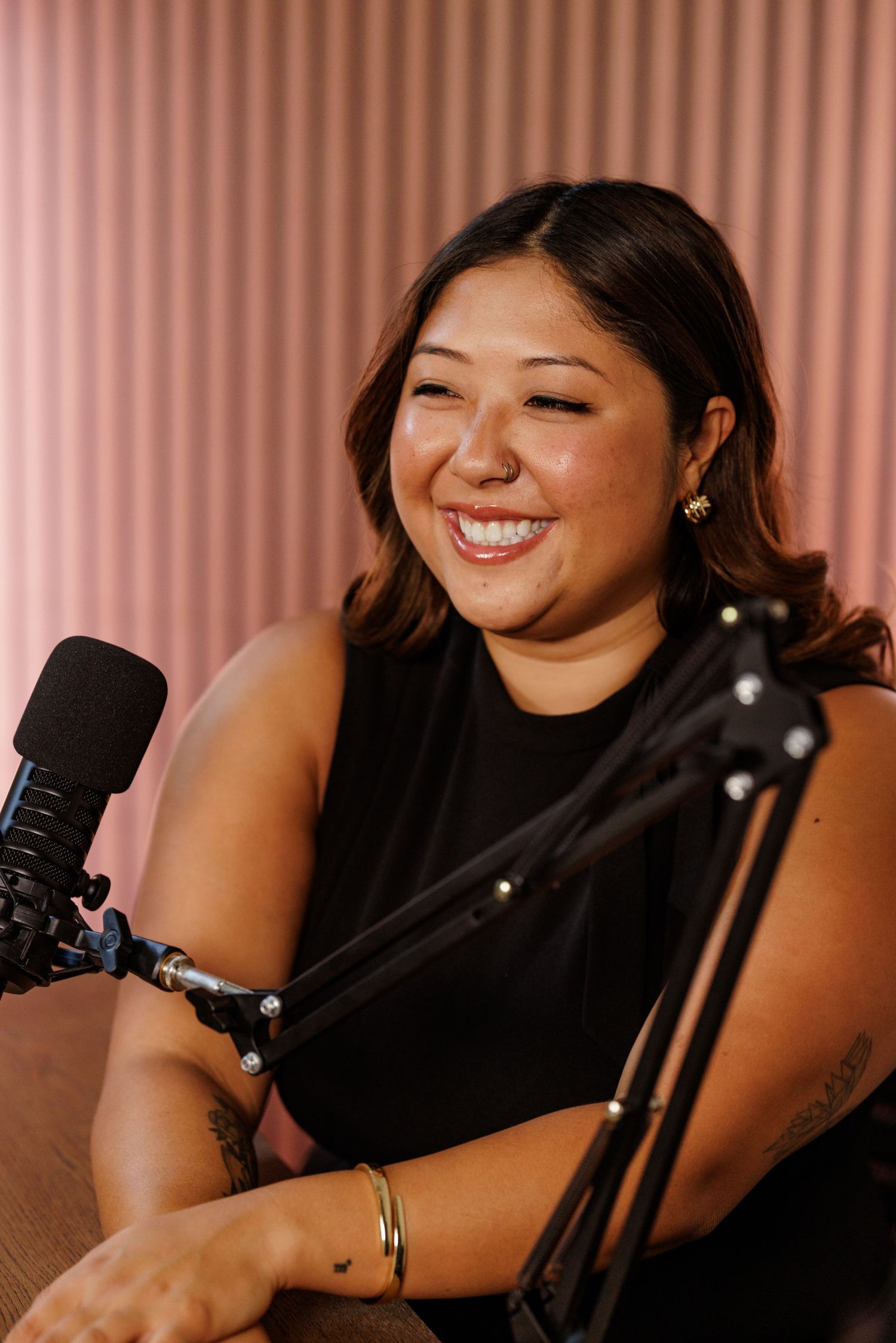 Smiling woman in black top recording a podcast with a microphone in a pink studio setting.