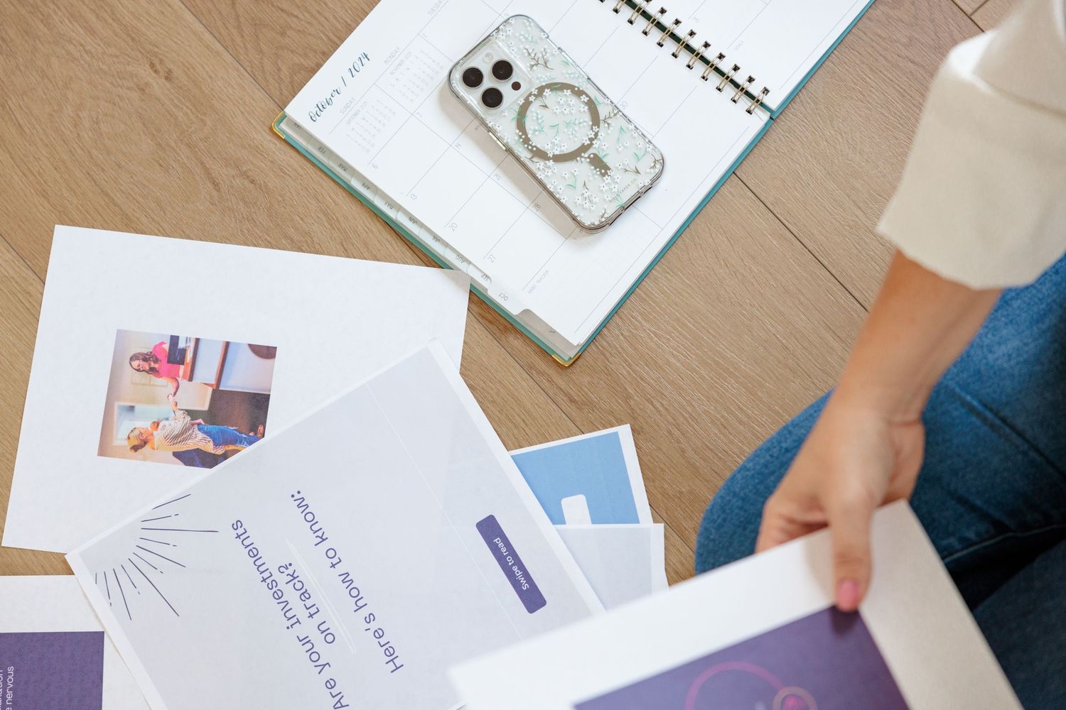 Person reviewing printed marketing materials and documents on a wooden floor beside a smartphone and open notebook.