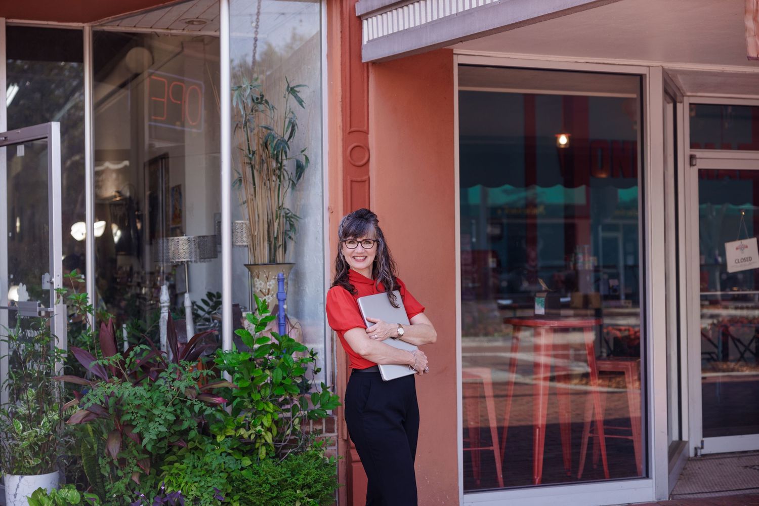 Smiling woman in red shirt holding a book stands at the entrance of a modern cafe or restaurant.