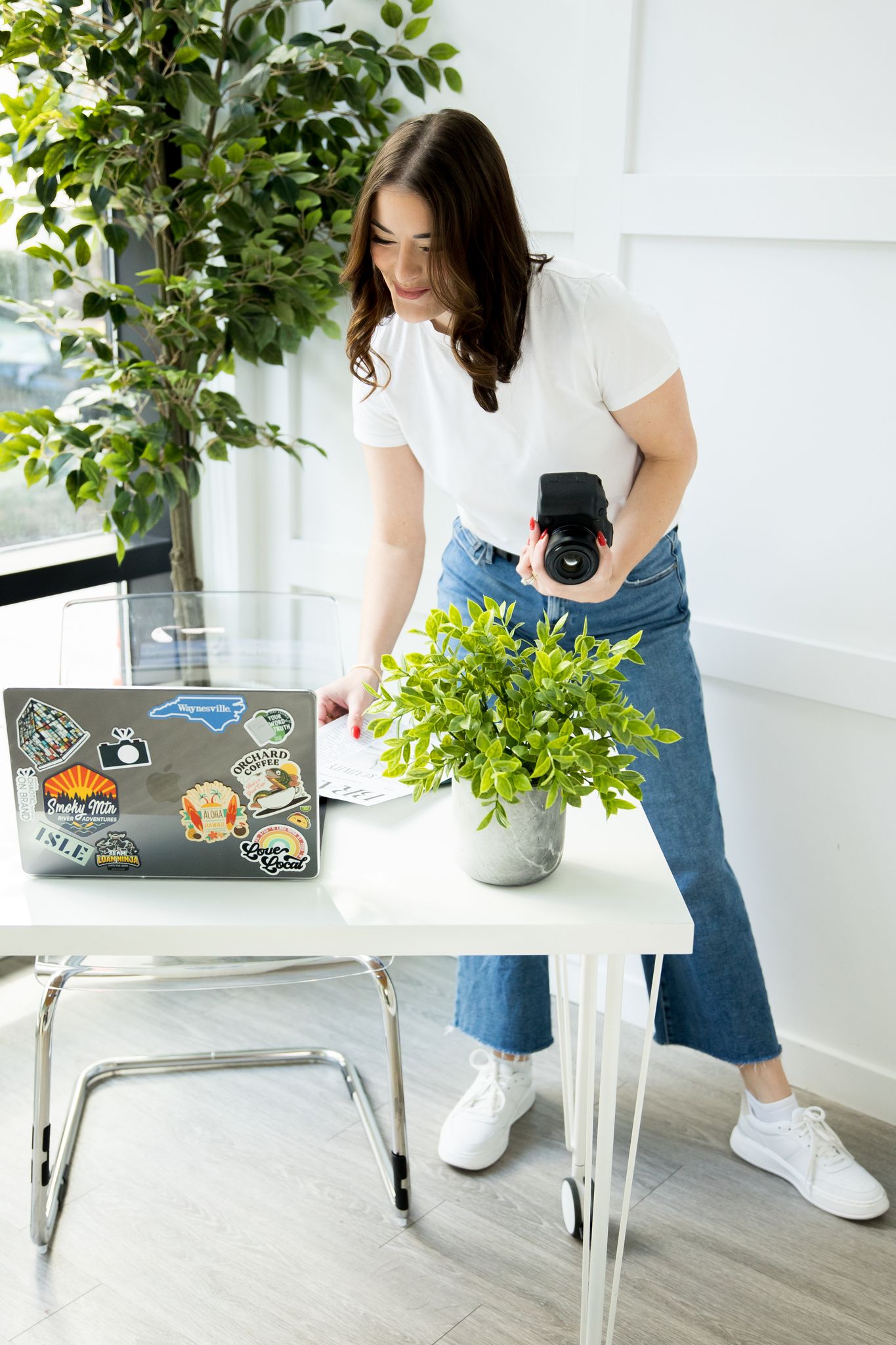 Woman in white tee and jeans holding camera near laptop with stickers and potted plant on white desk.