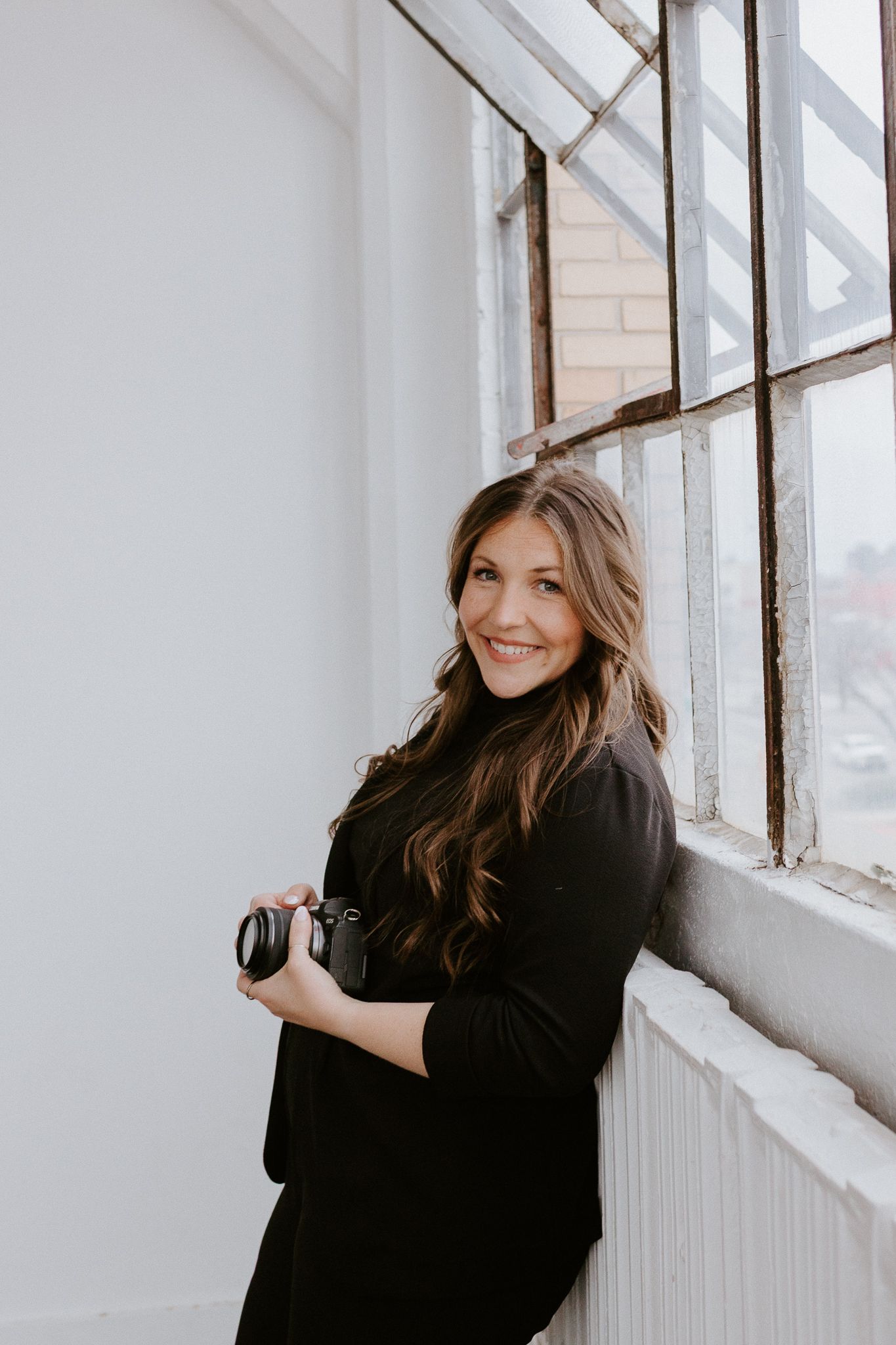 Professional photographer in black standing by bright window with camera during indoor photoshoot.