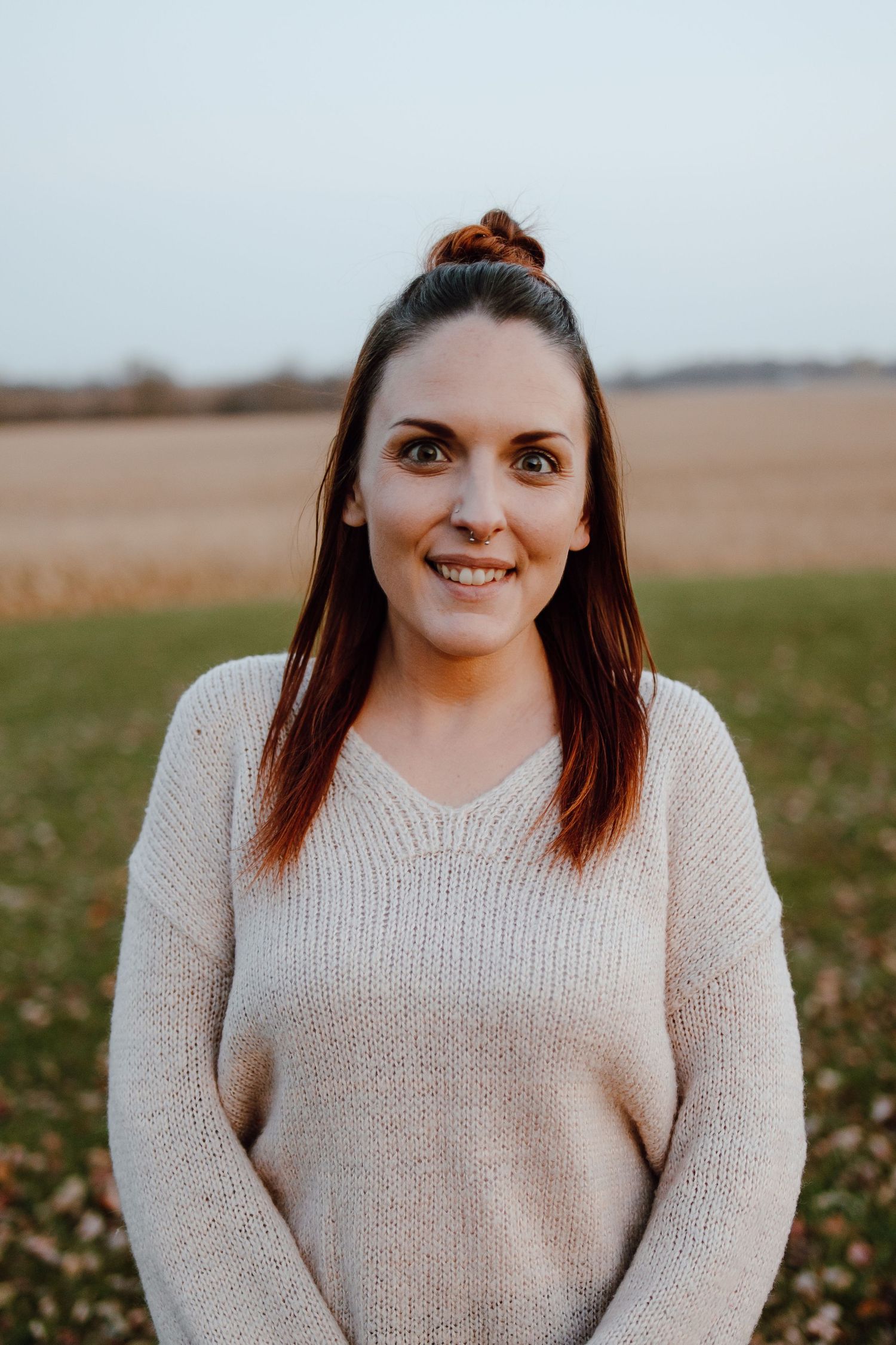 A person wearing a beige knit sweater smiles warmly while standing outdoors in an autumn field.