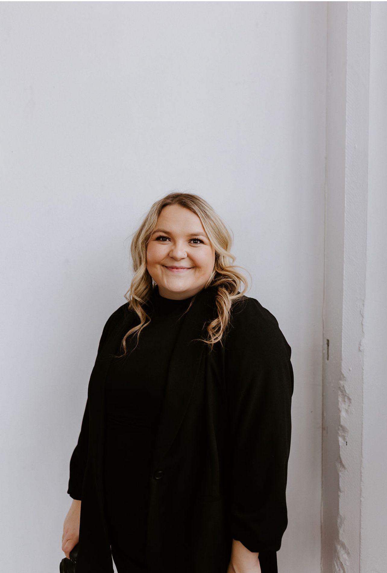 Person wearing a black turtleneck sweater smiling against a white wall in minimalist portrait setting.