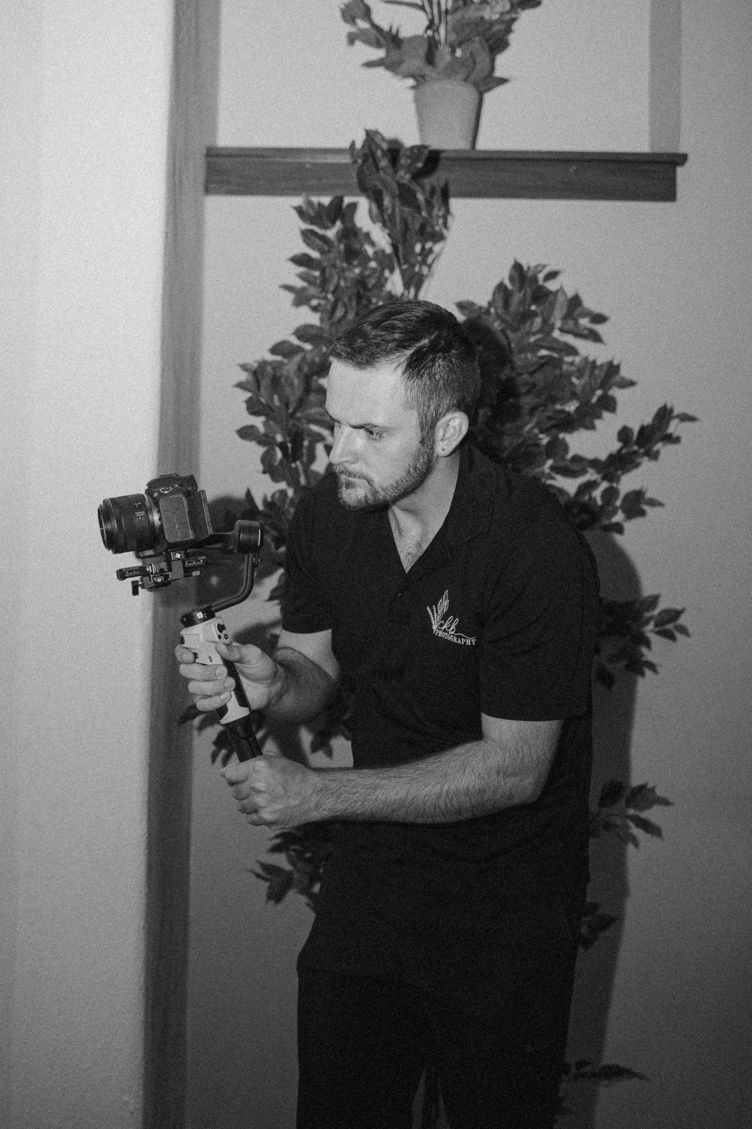Man holding a camera gimbal stabilizer outdoors, black and white photography with plants in background.