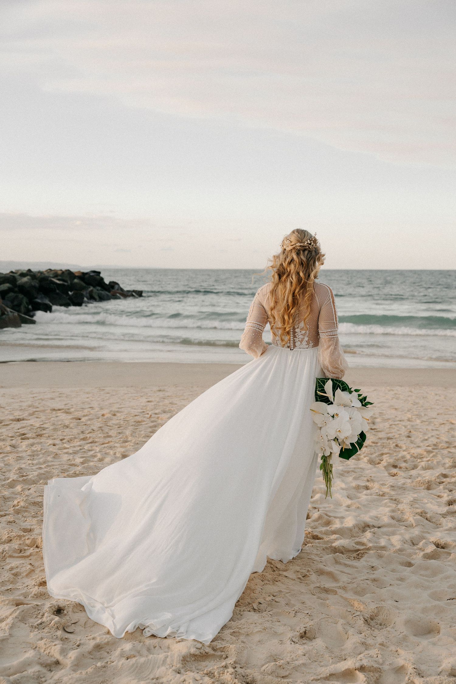 Bride in white flowing gown holding bouquet stands on sandy beach with ocean waves and rocks in background.