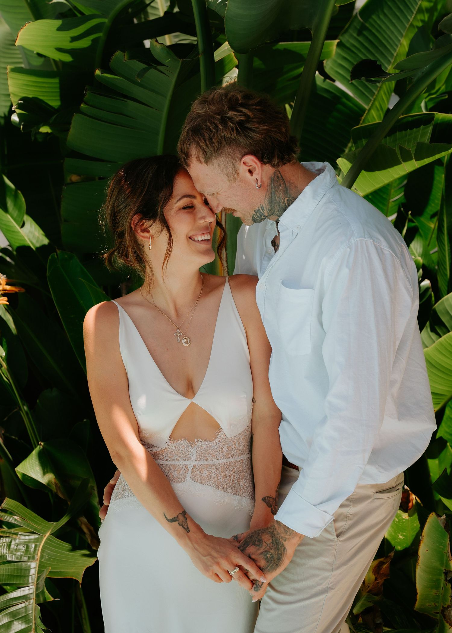 A smiling bride in a white dress and groom in a white shirt share a romantic moment surrounded by tropical greenery.