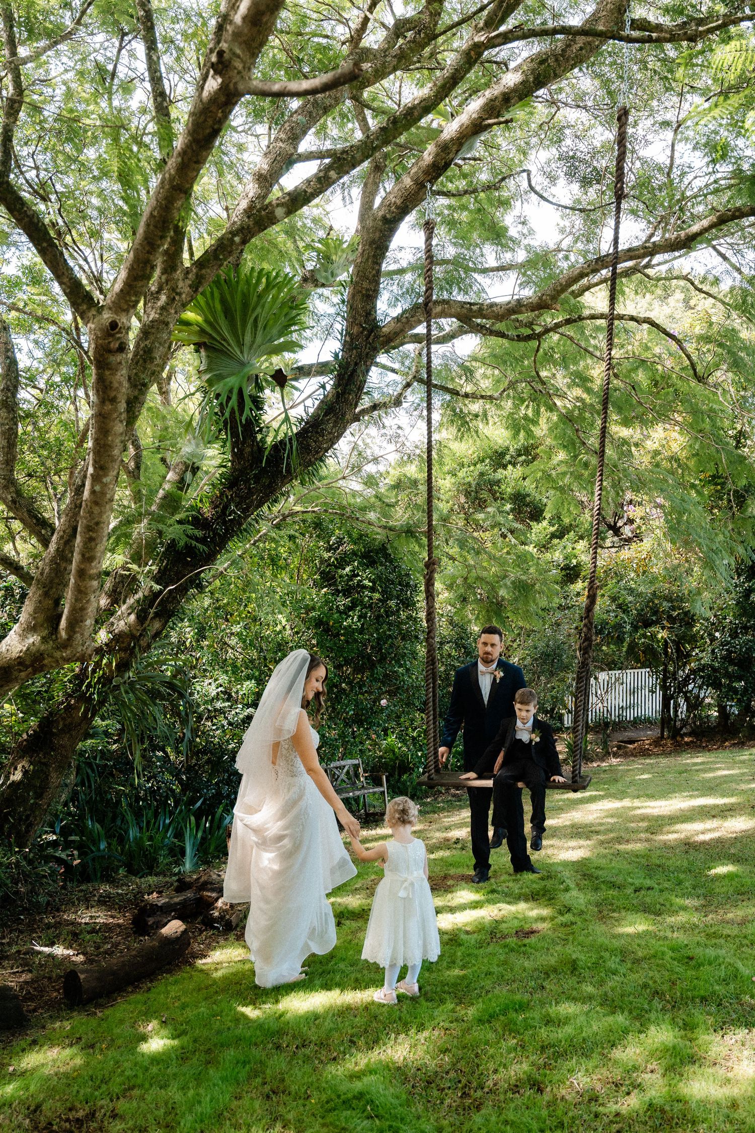 Bride in white gown holds flower girl's hand as groom in black suit approaches under lush garden trees.