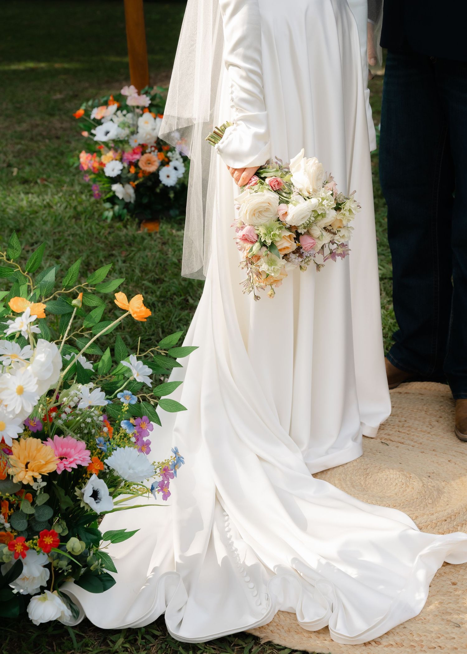 Bride in white wedding gown holding pastel bouquet, surrounded by colorful floral arrangements at outdoor ceremony.