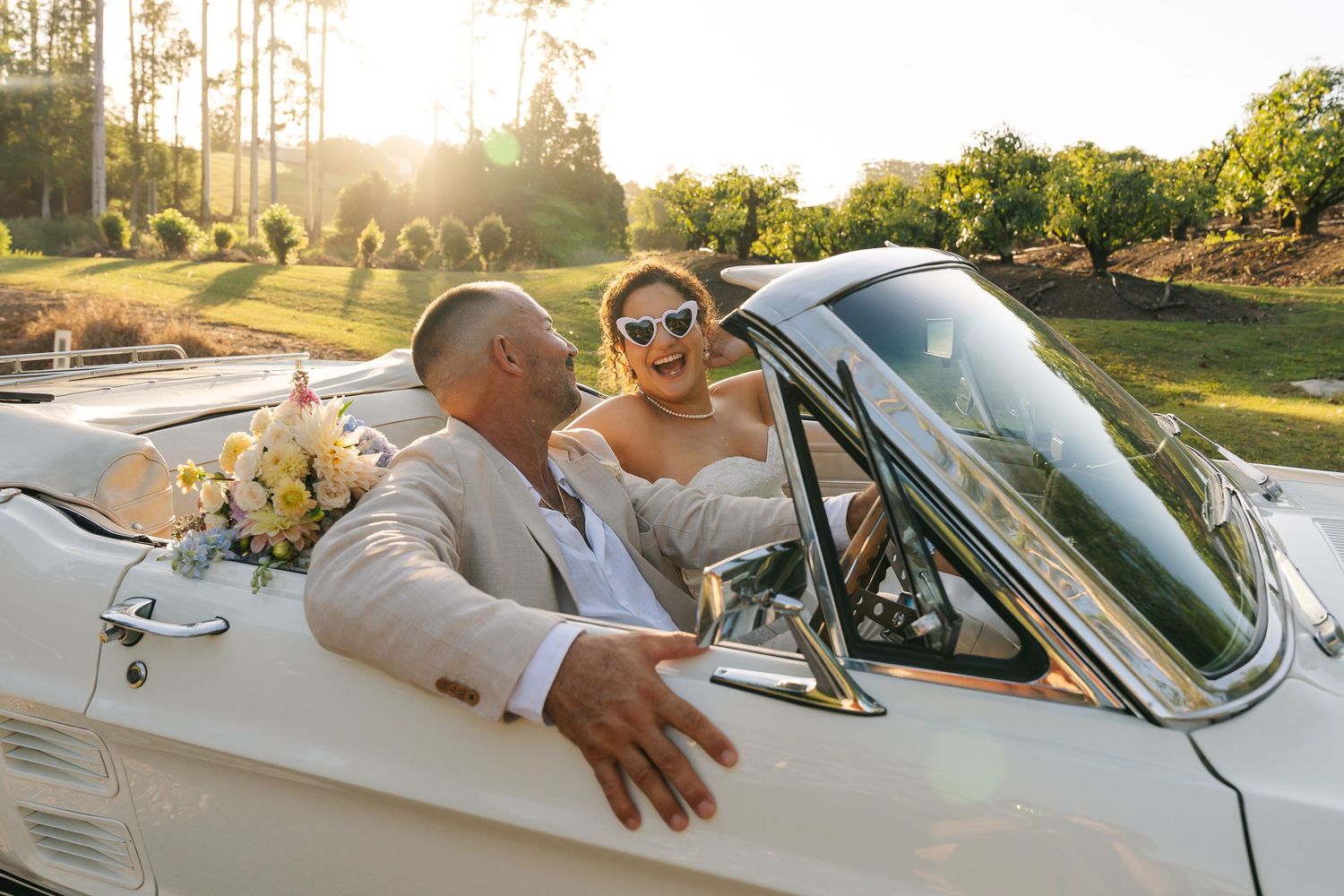 Bride and groom in wedding attire sitting in a white vintage convertible car at sunset.