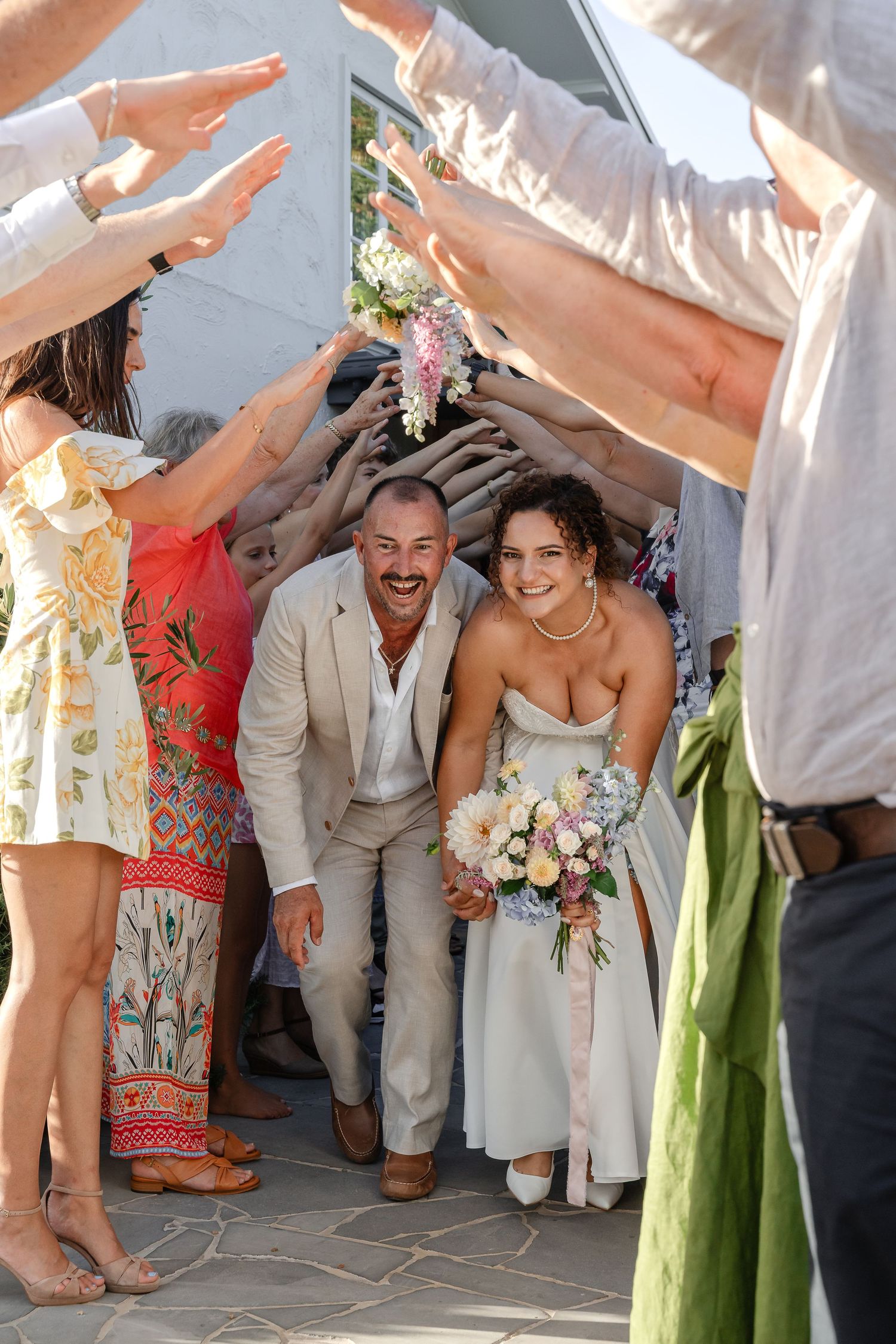 Joyful newlyweds walk through tunnel of raised hands formed by wedding guests after ceremony.