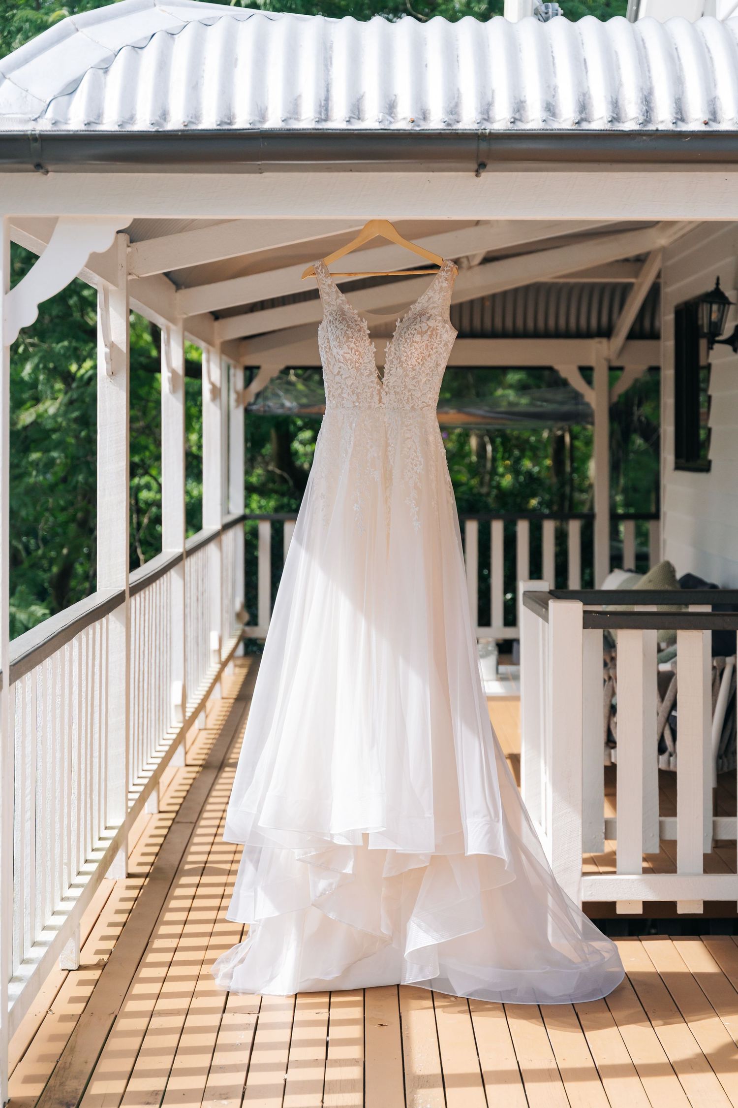 Elegant white lace A-line wedding dress hanging on wooden hanger on sunlit outdoor veranda with white railings.