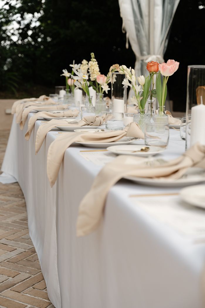 Elegant outdoor wedding reception table with white linen, beige napkins, and fresh tulip floral arrangements.