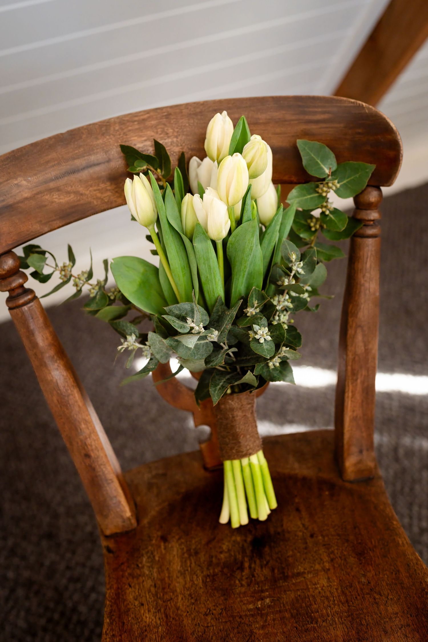 White tulip bridal bouquet with green foliage tied with twine, resting on a rustic wooden chair.