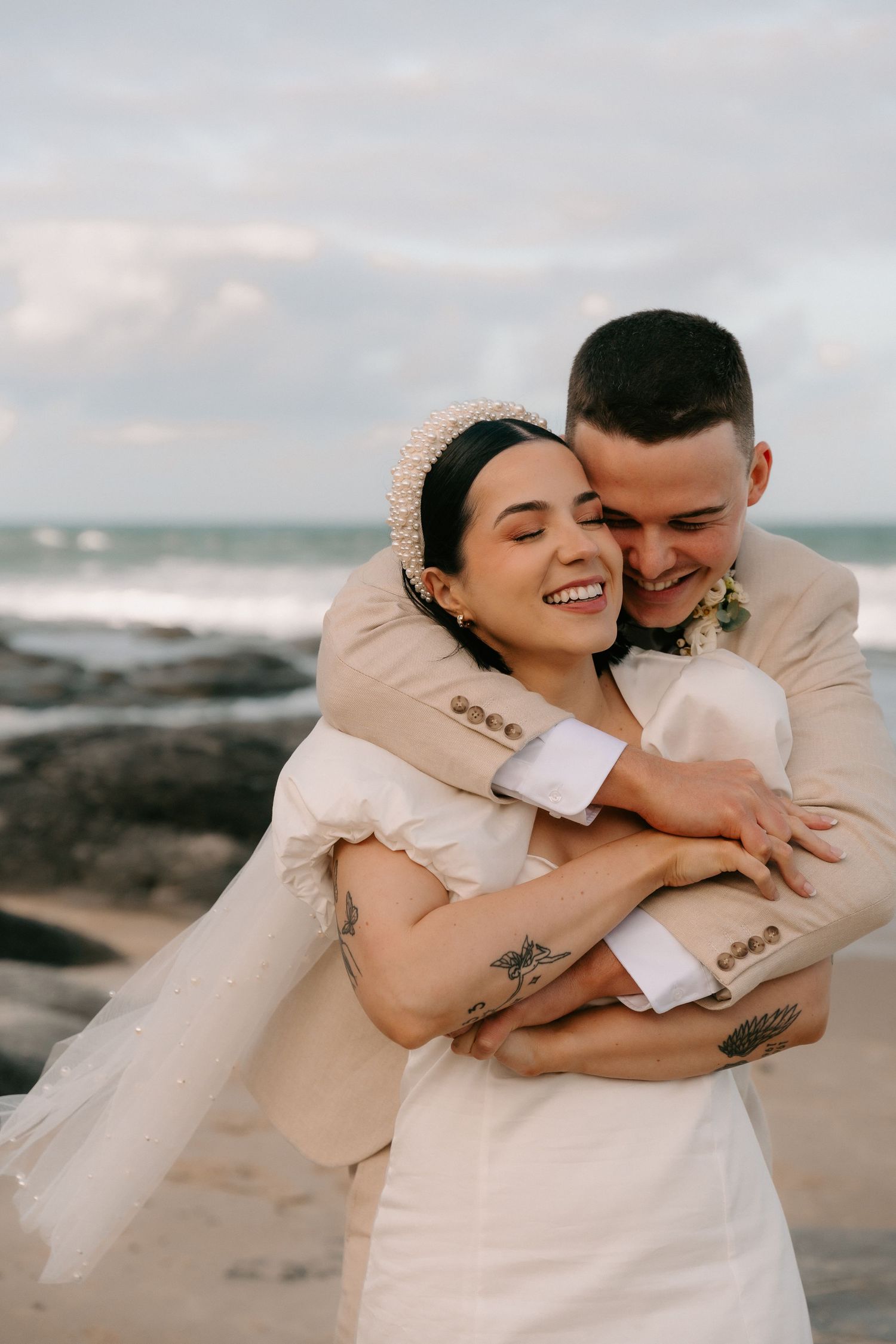 Joyful wedding couple embracing on rocky beach during sunset.