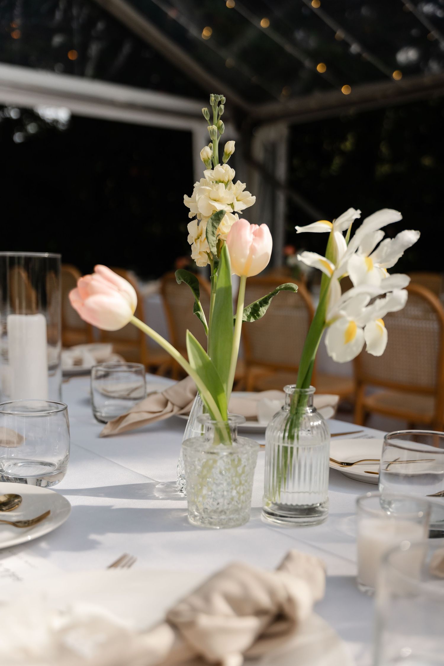 Elegant wedding table setting with pink tulips and white iris flowers in glass vases on a white tablecloth.