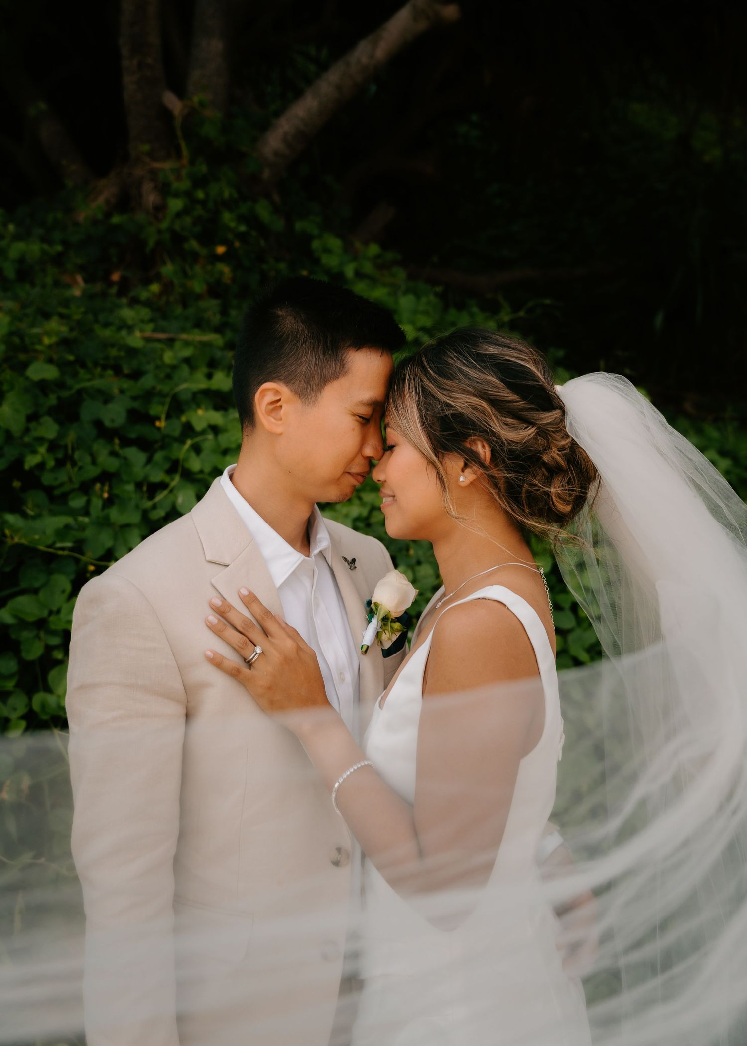 A bride and groom share an intimate forehead touch, her veil flowing around them, surrounded by lush greenery.