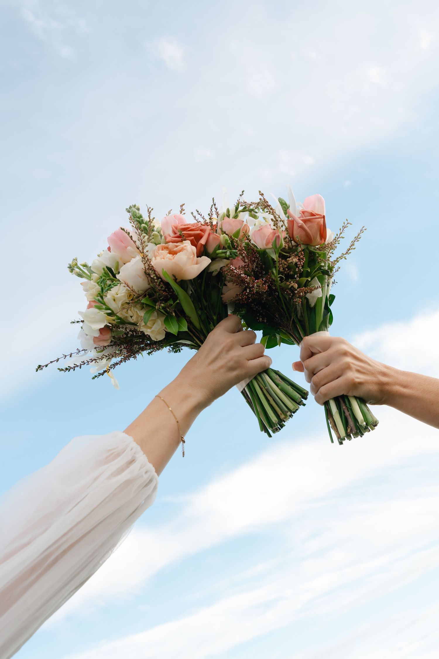 Two hands holding a beautiful bridal bouquet of pink roses and wildflowers against a blue sky.