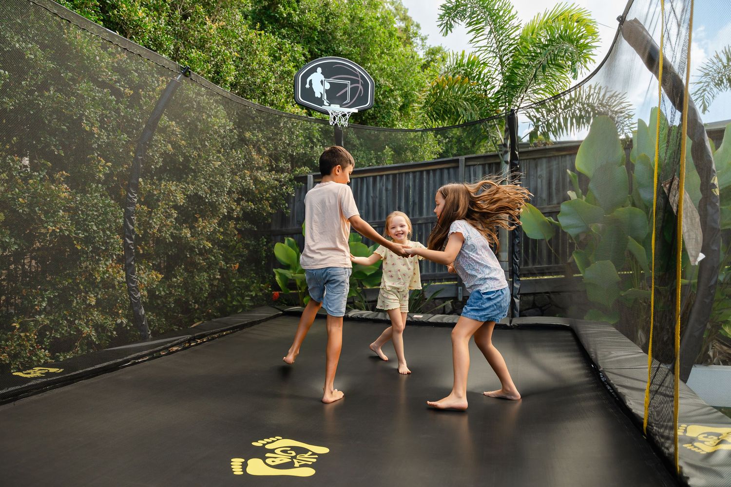 Three children playing and holding hands on a backyard trampoline with a basketball hoop and green hedge backdrop.