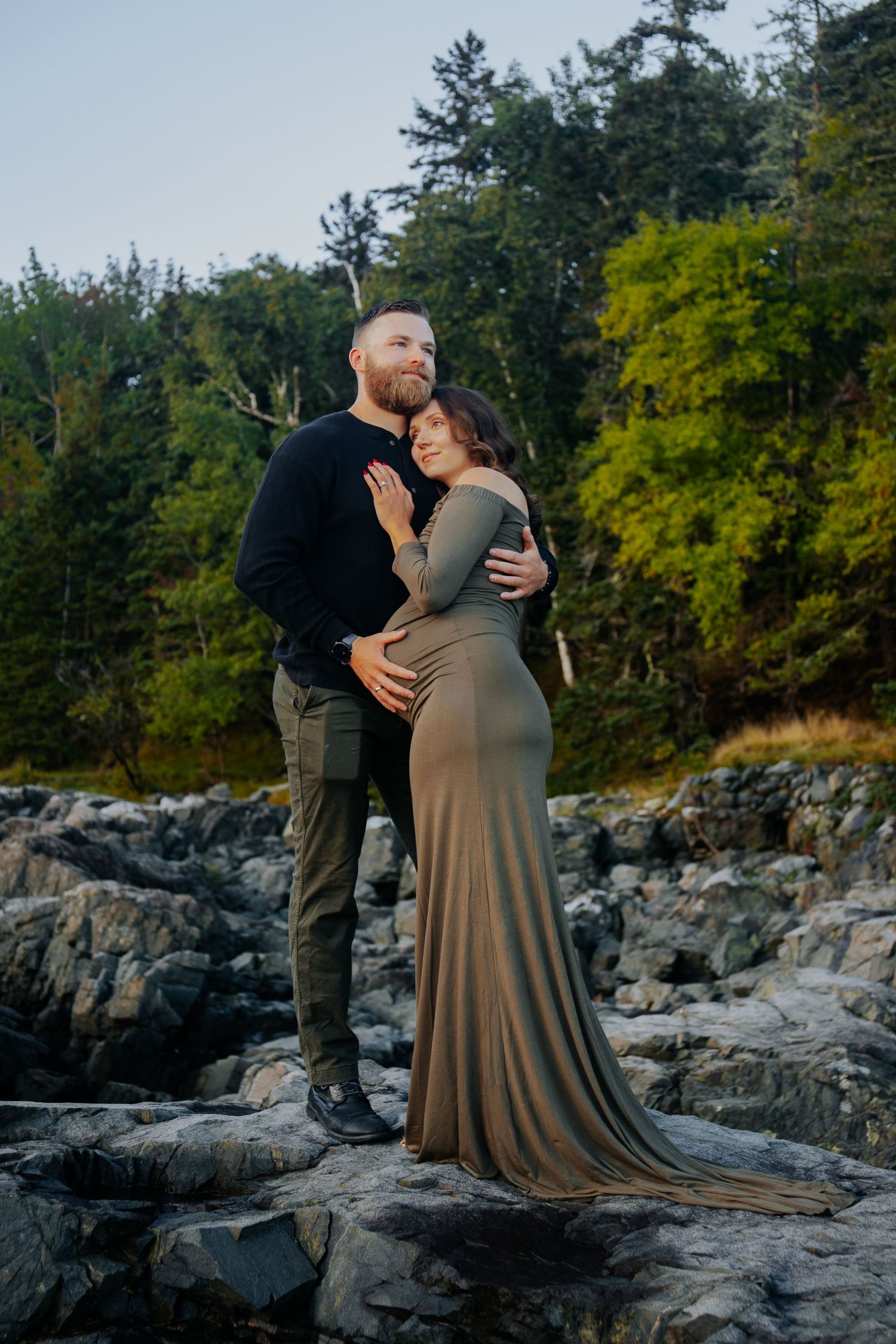 A couple embraces on rocky coastal terrain surrounded by green trees at golden hour.