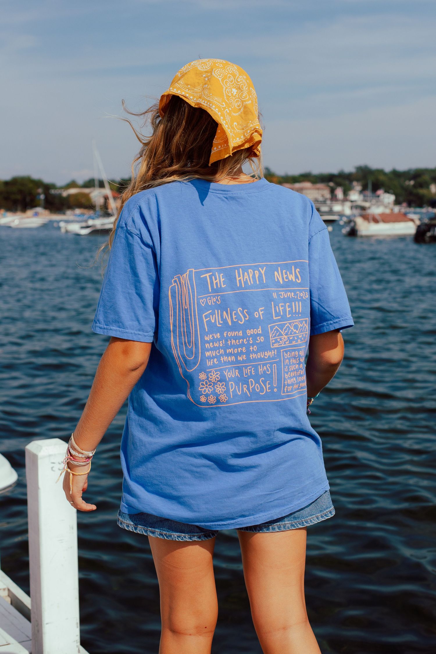 Light blue t-shirt with text design shown on boat deck against coastal water backdrop.