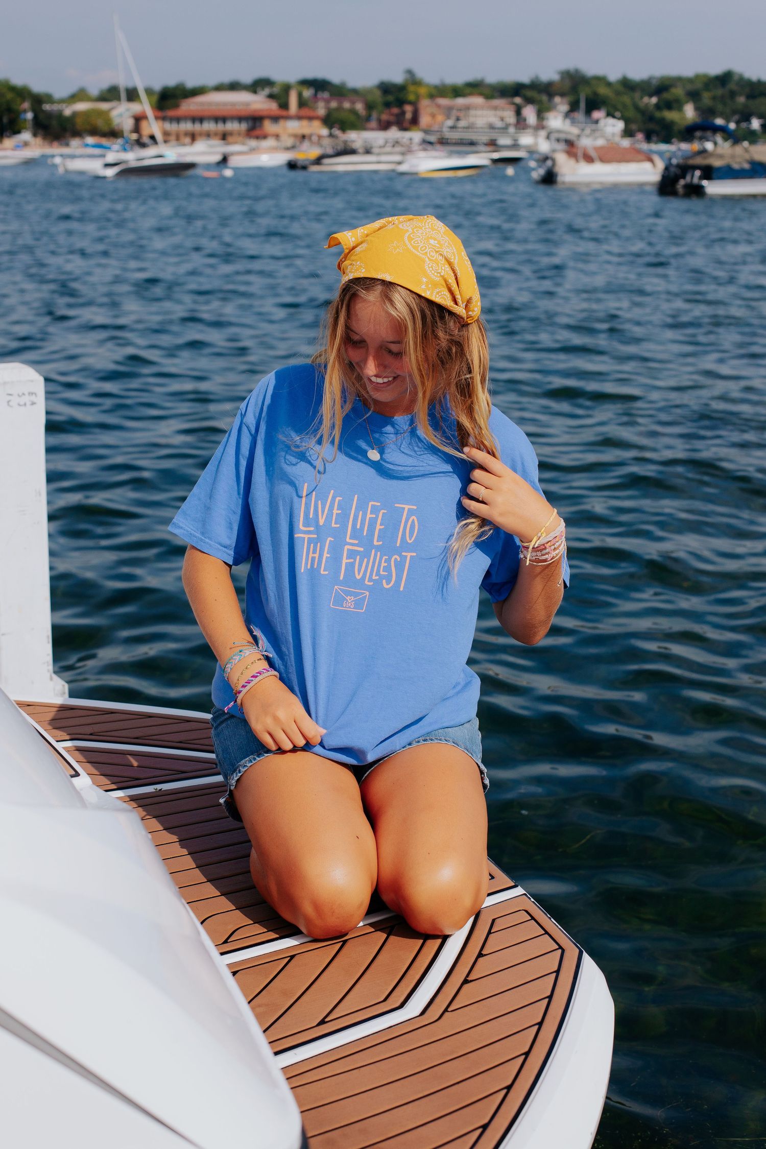 Person relaxing on wooden boat deck wearing blue t-shirt and yellow hat near marina.
