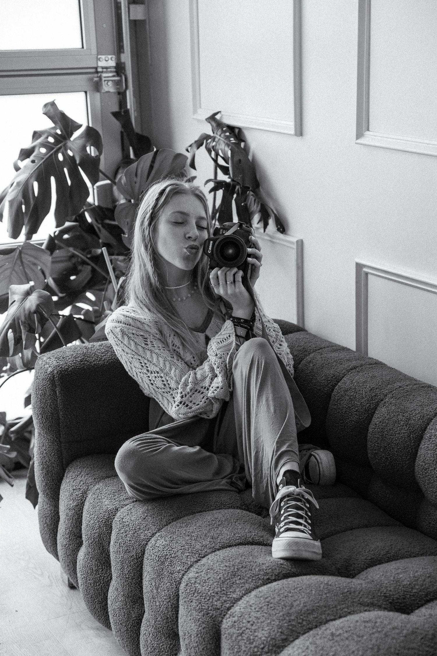 Black and white photo of person with camera sitting on tufted couch near monstera plant.