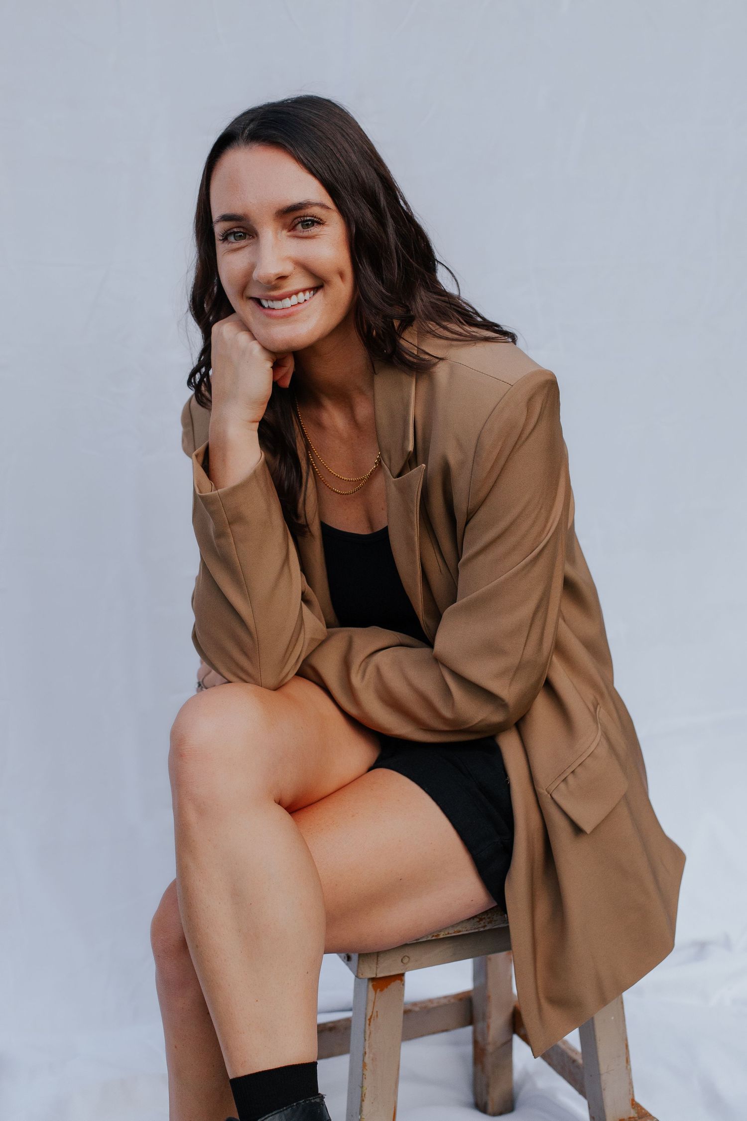 Person in tan blazer and black top seated on wooden stool during photo shoot.