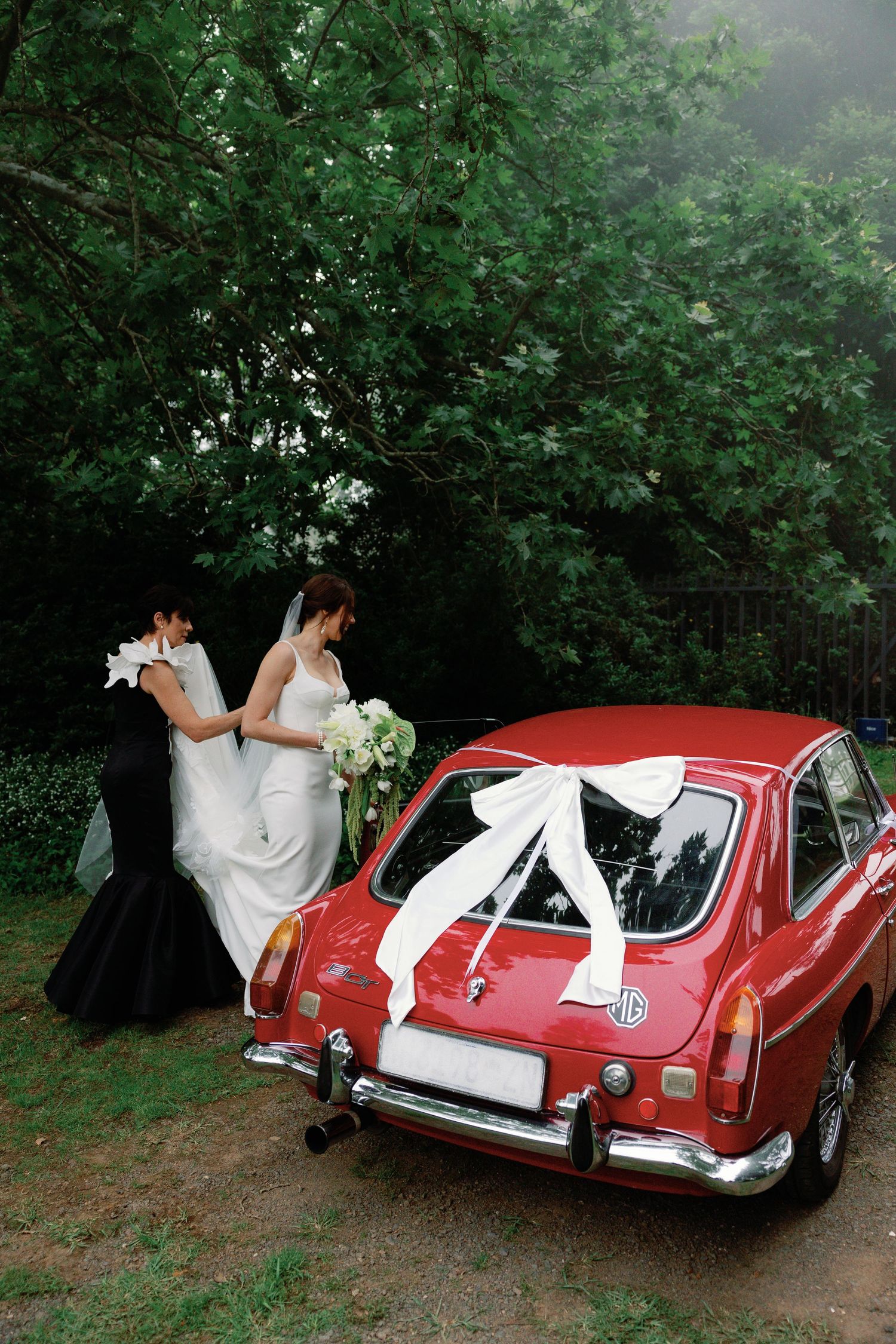 A wedding couple poses with a classic red MGB GT sports car in a wooded setting.