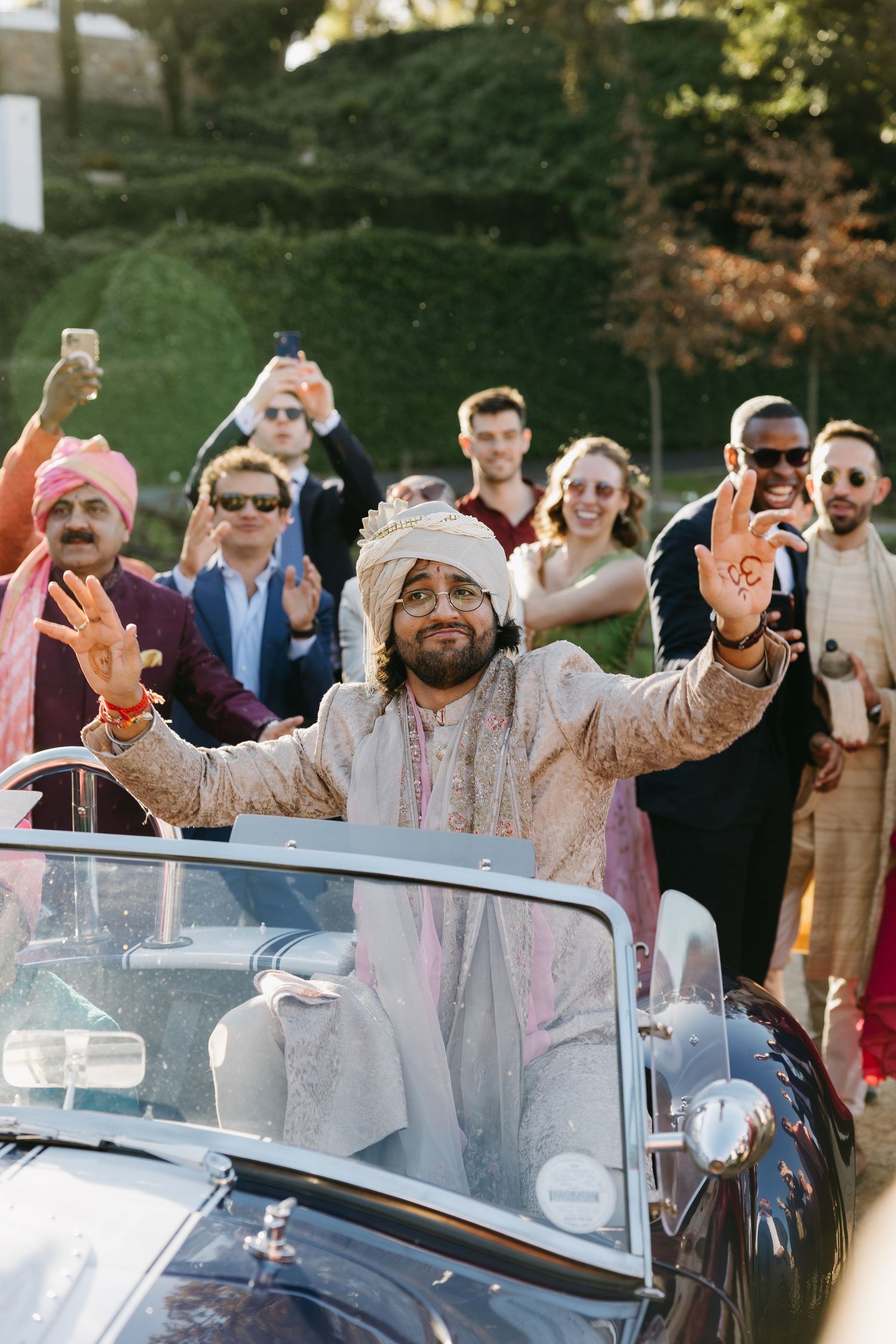 Wedding guests celebrate joyfully around a vintage car during an outdoor celebration.