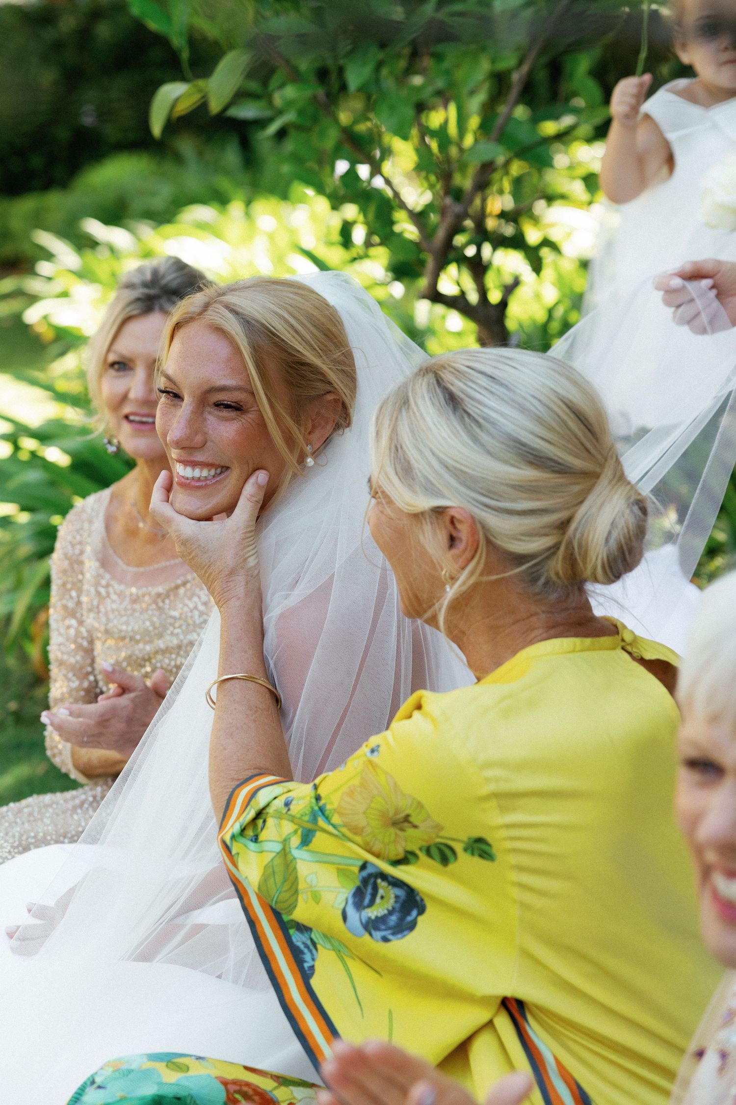 Smiling bride in veil with wedding guests in garden, woman in yellow floral dress touching her face.