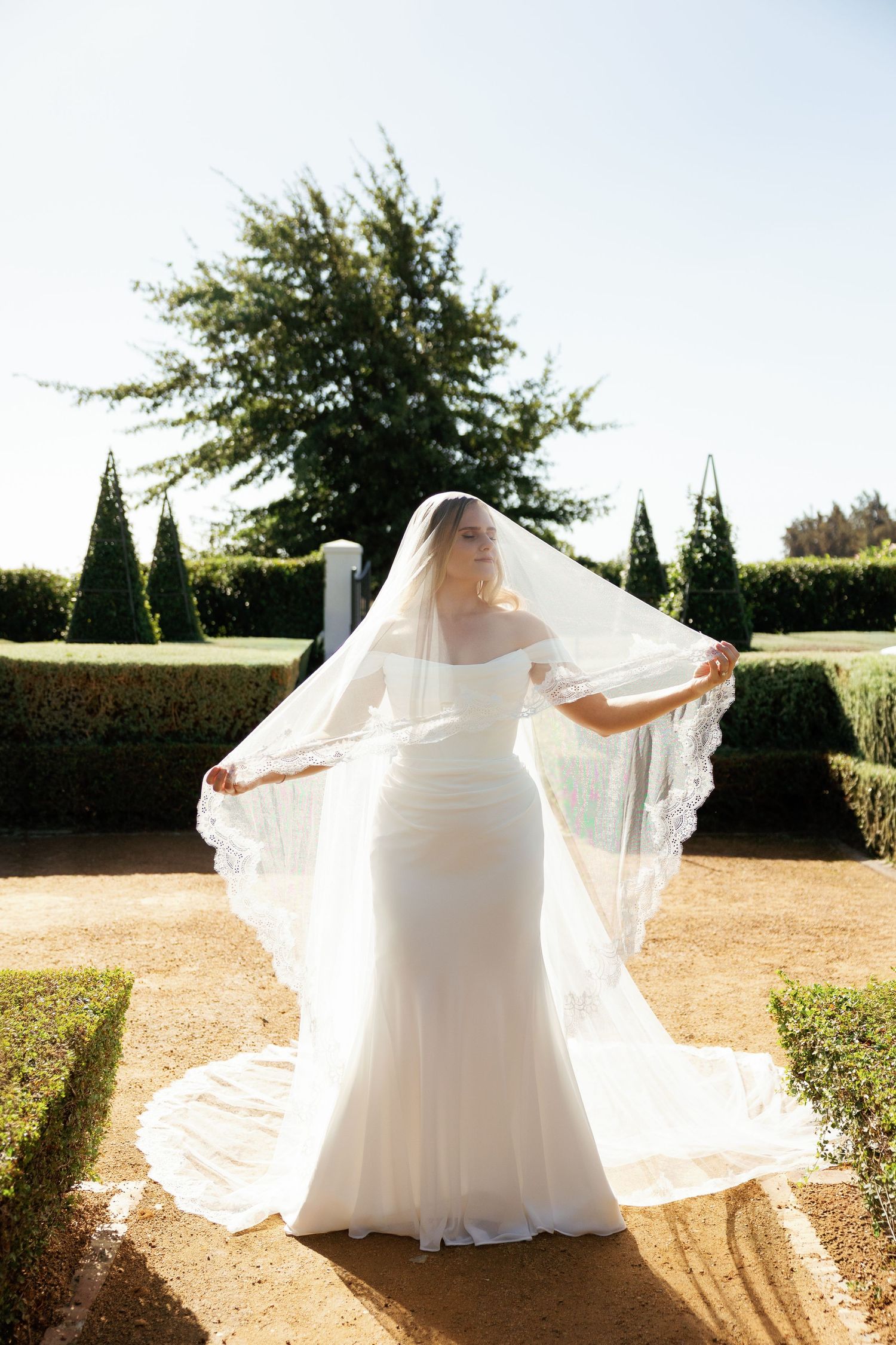 Bride in white gown lifting lace veil in formal garden with manicured hedges and cypress trees.