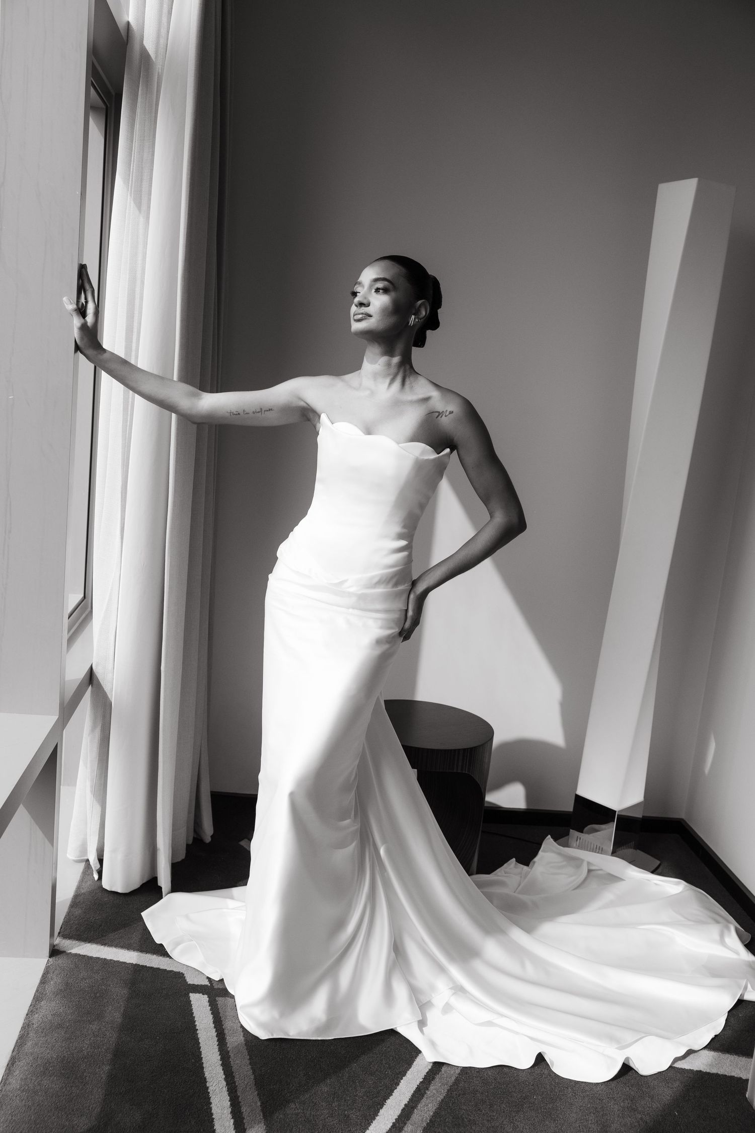 Black and white photo of a bride in a strapless white gown with a train, posing elegantly by a window.