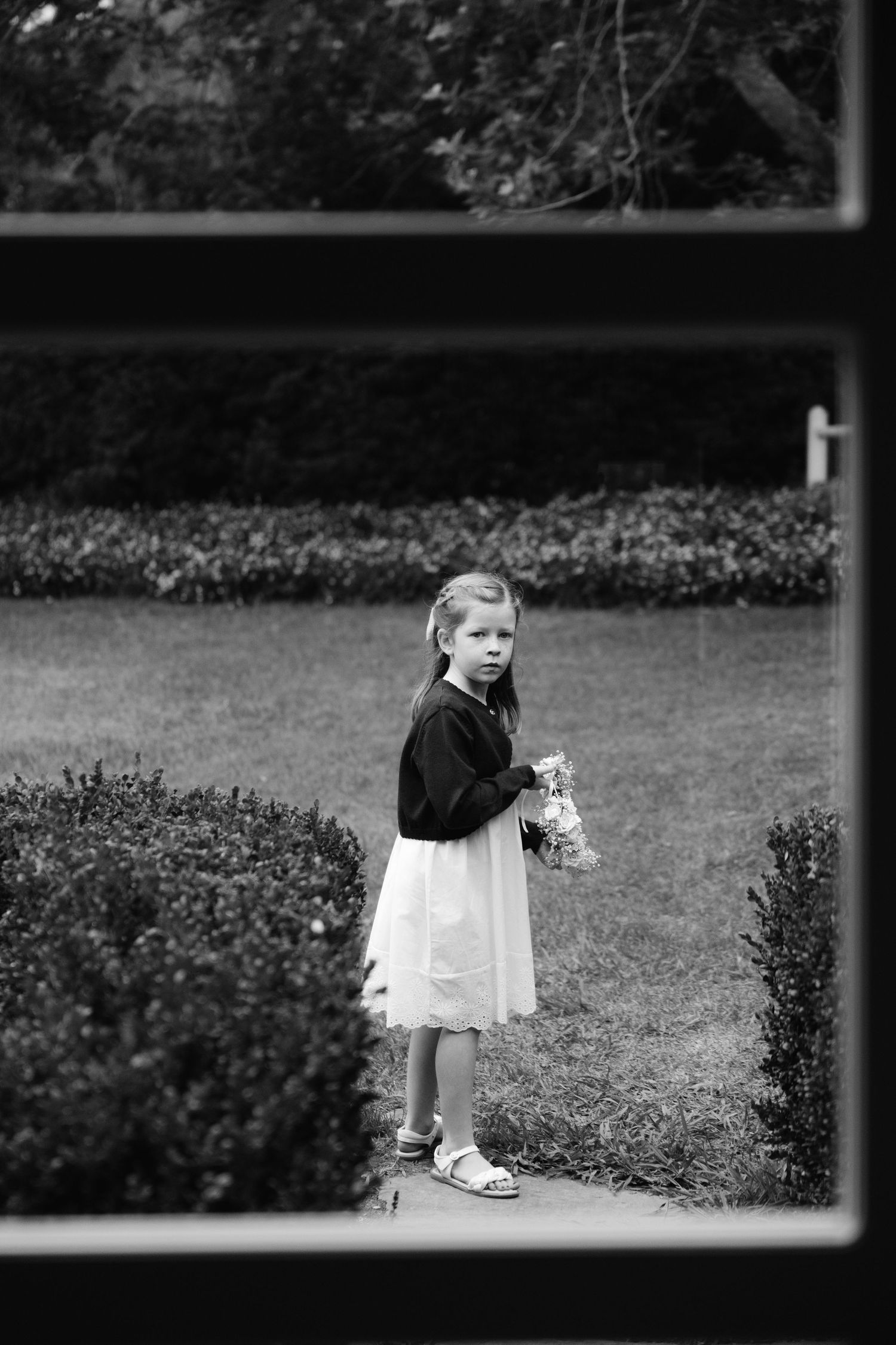 Black and white photo of a young girl in white skirt holding flowers, standing on a garden path surrounded by hedges.