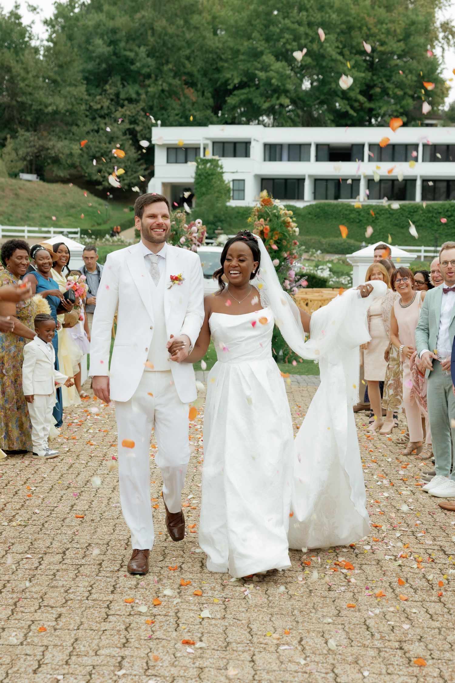 Bride and groom walk through flower petal confetti shower, surrounded by guests at outdoor wedding ceremony.
