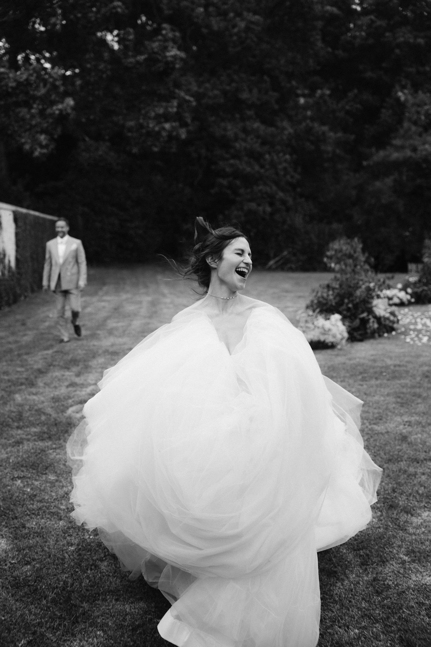 Black and white photo of a laughing bride twirling in a voluminous wedding gown outdoors with a guest approaching in background.