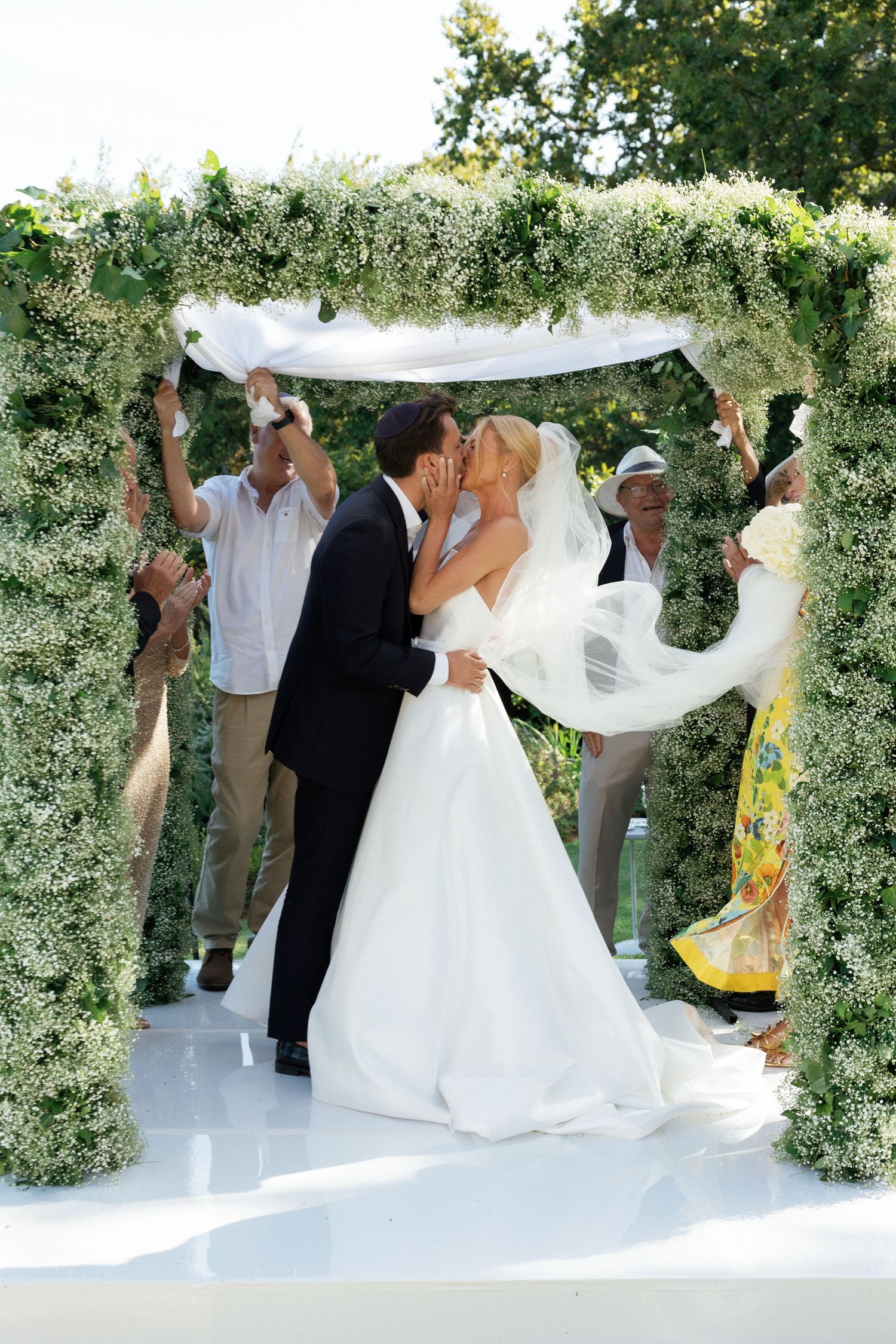 Bride and groom share first kiss under floral wedding arch outdoors, surrounded by guests celebrating.