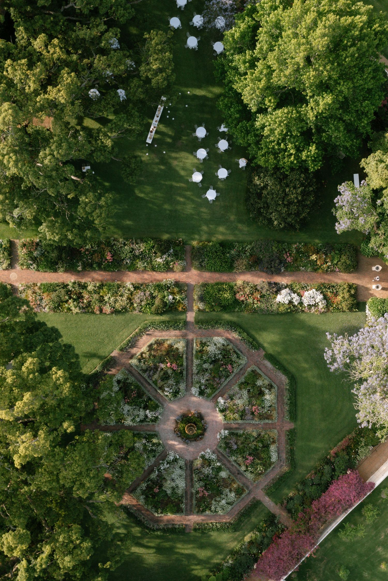 Aerial view of a formal garden with winding brick pathways and geometric patterns surrounded by lush green trees.