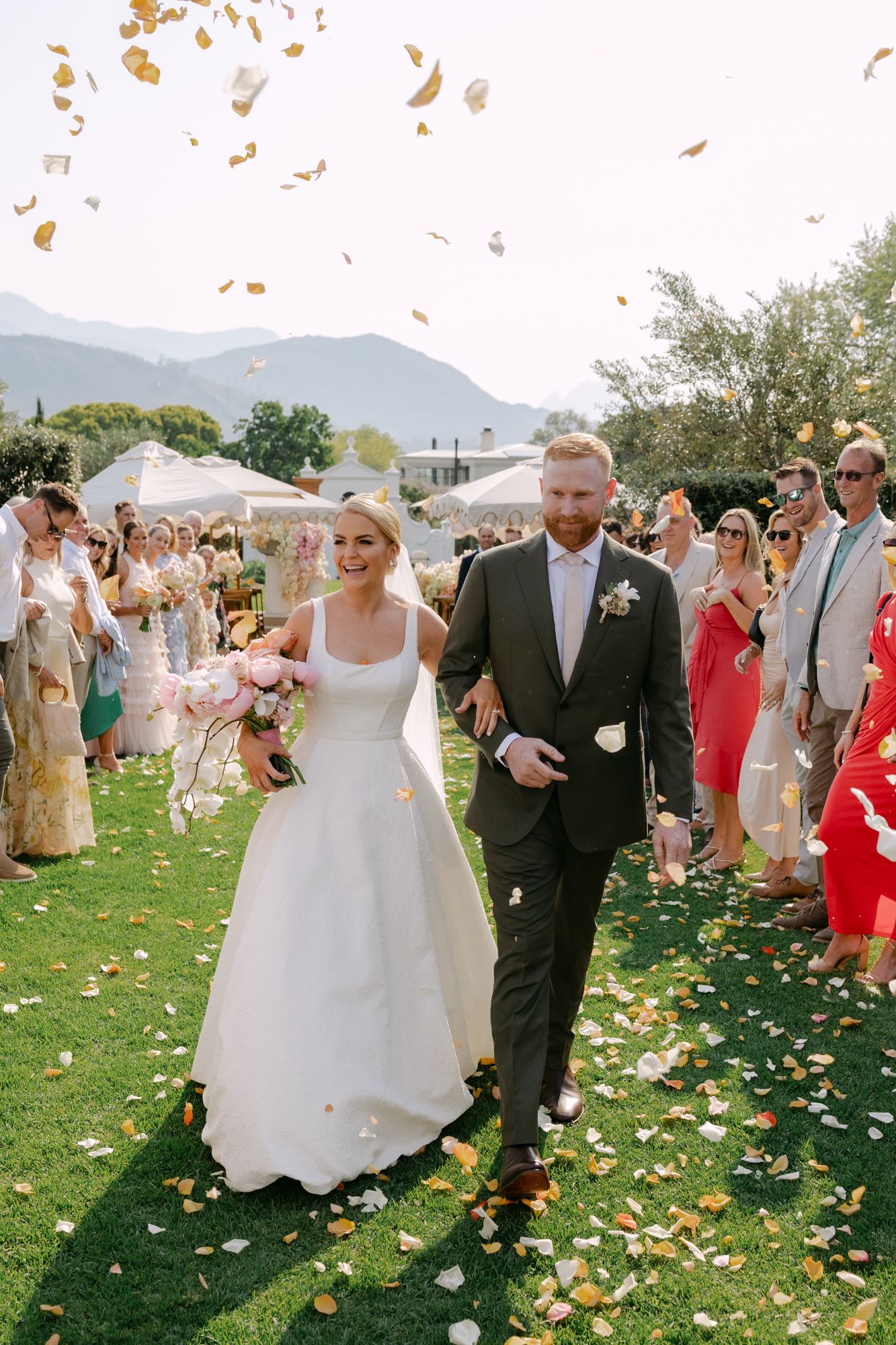 Wedding guests shower newlyweds with golden leaves on a grassy lawn with mountains in the background.