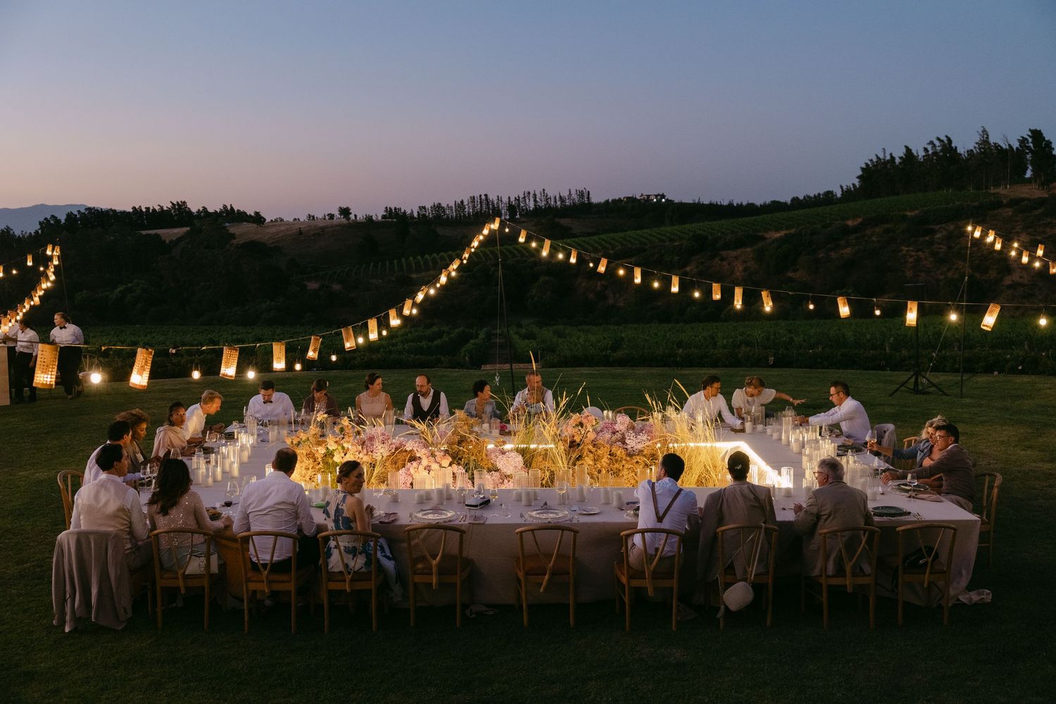 String lights illuminate an elegant outdoor wedding reception setup with white tables and chairs at dusk.
