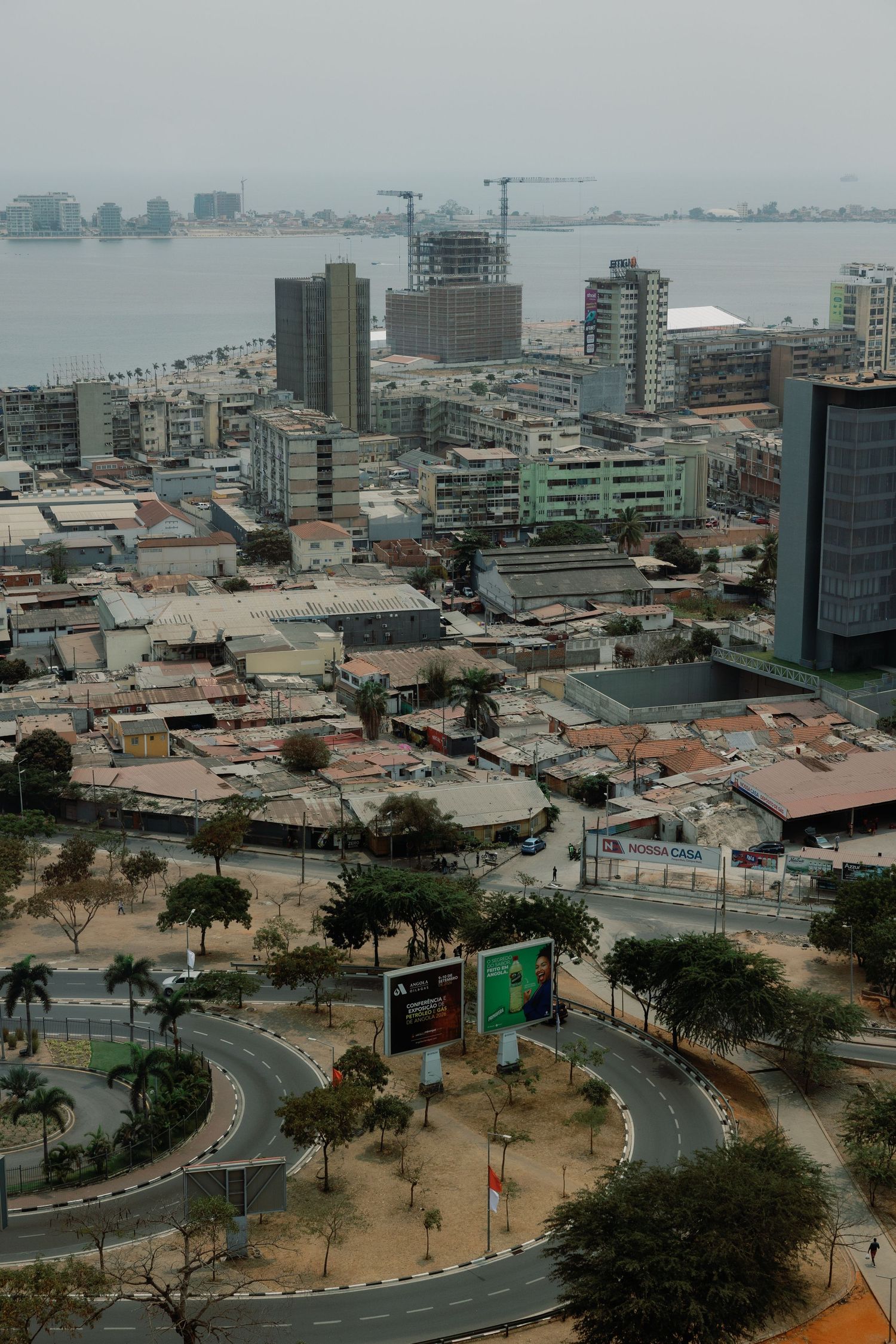 Aerial view of a coastal African city with winding roads, palm trees, urban buildings, and a bay visible in the background.