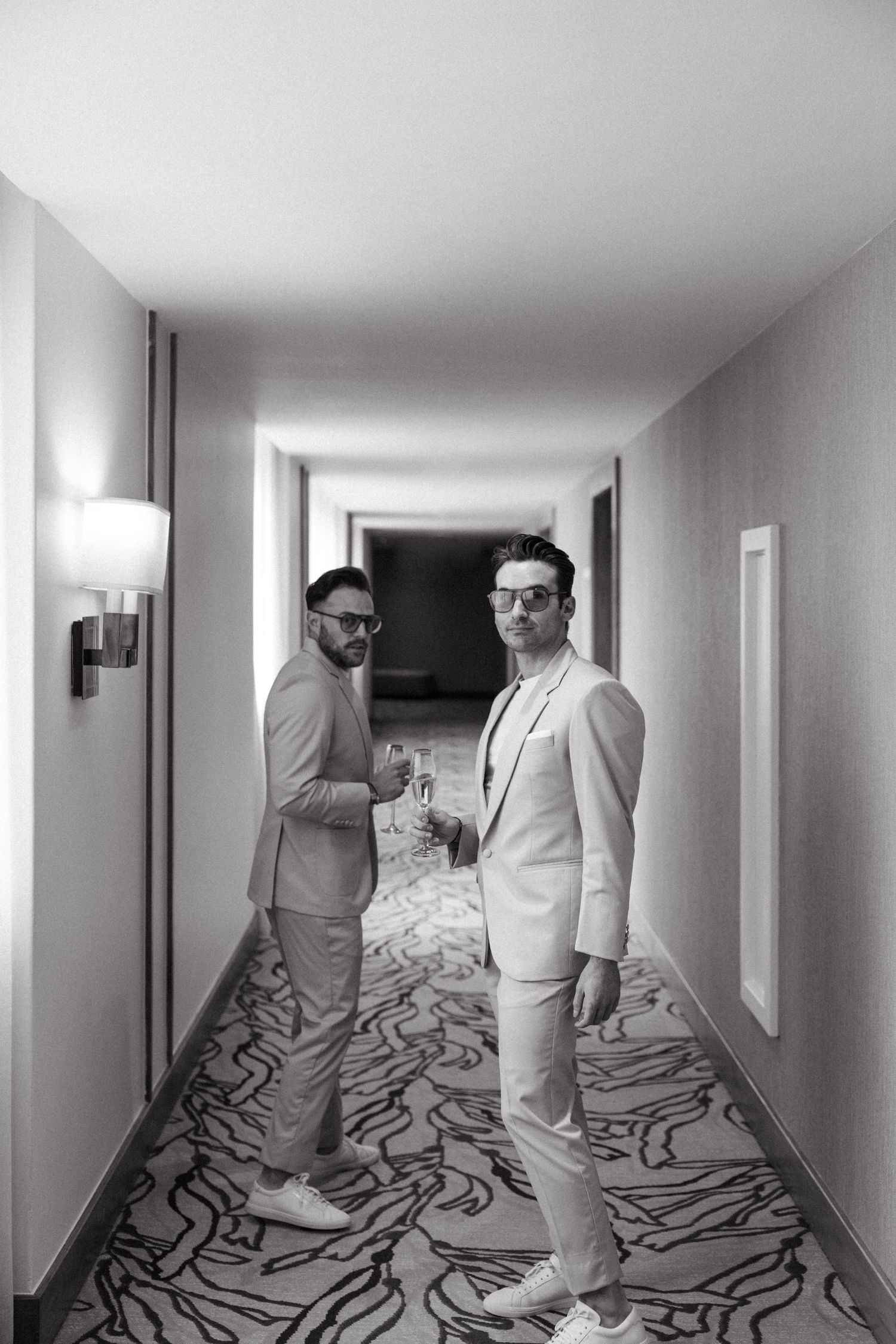 Two men in light suits and sunglasses stand in a hotel hallway, holding drinks in a black and white photo.