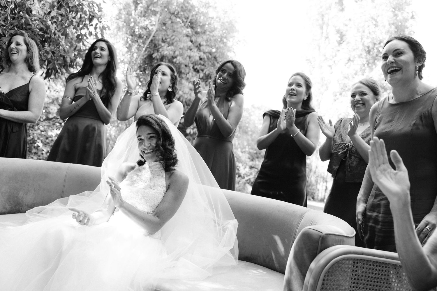 Black and white photo of a bride in a white gown seated, surrounded by smiling bridesmaids in dark dresses clapping outdoors.