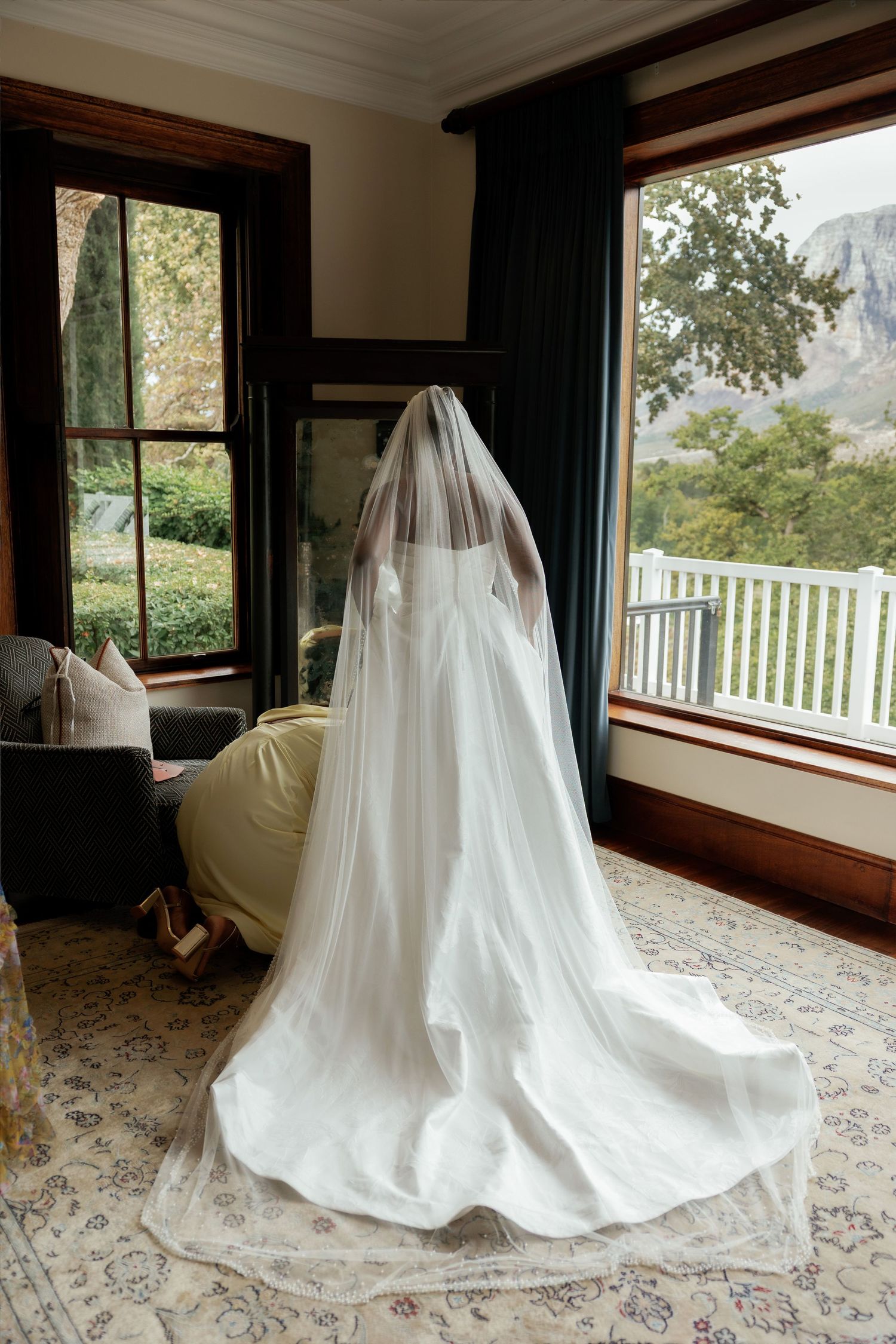 Bride in white wedding gown and long veil stands by open window with mountain view on her wedding day.