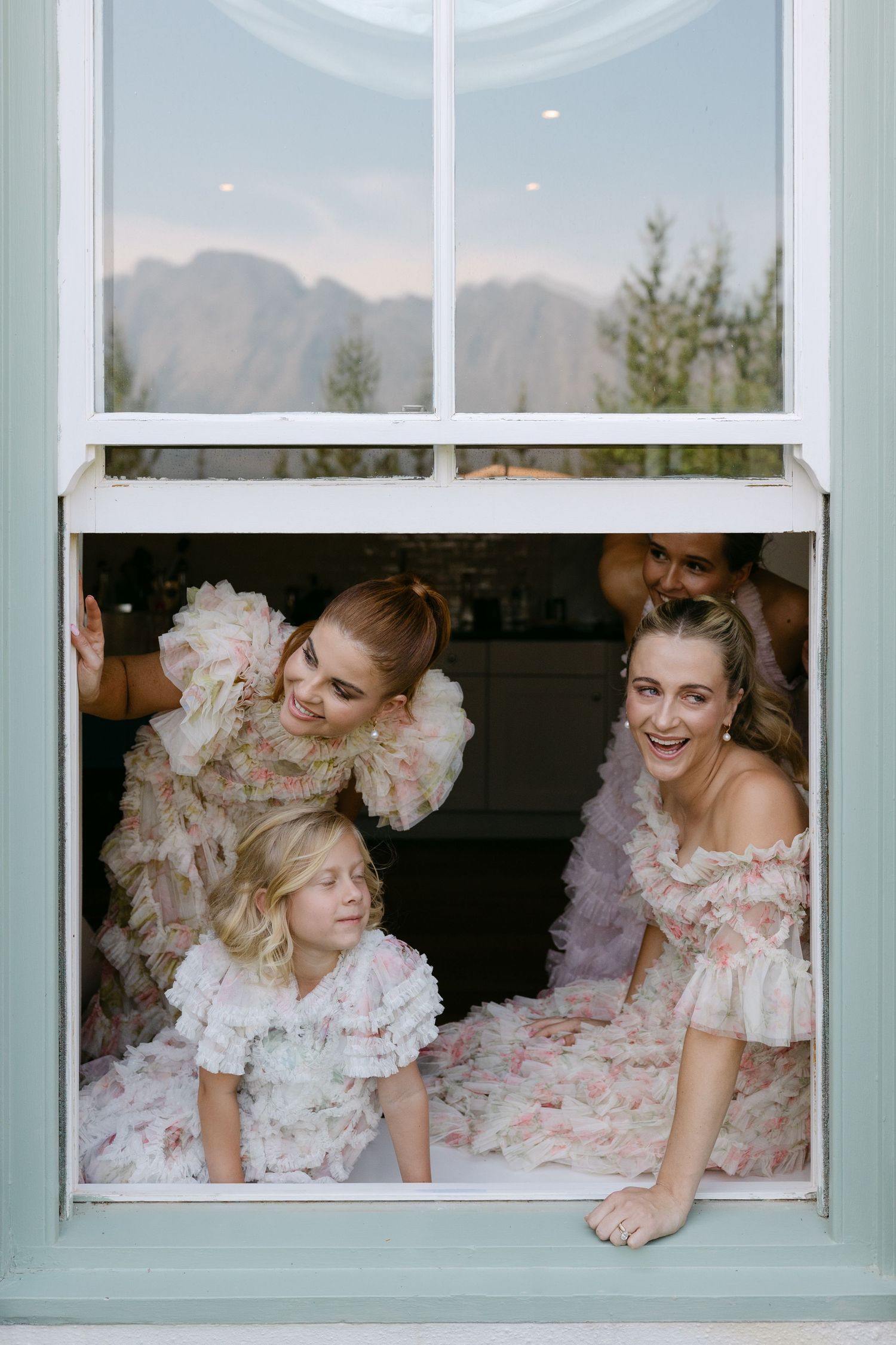 Three people in pink ruffled dresses peek playfully through a mint green window frame at a wedding celebration.