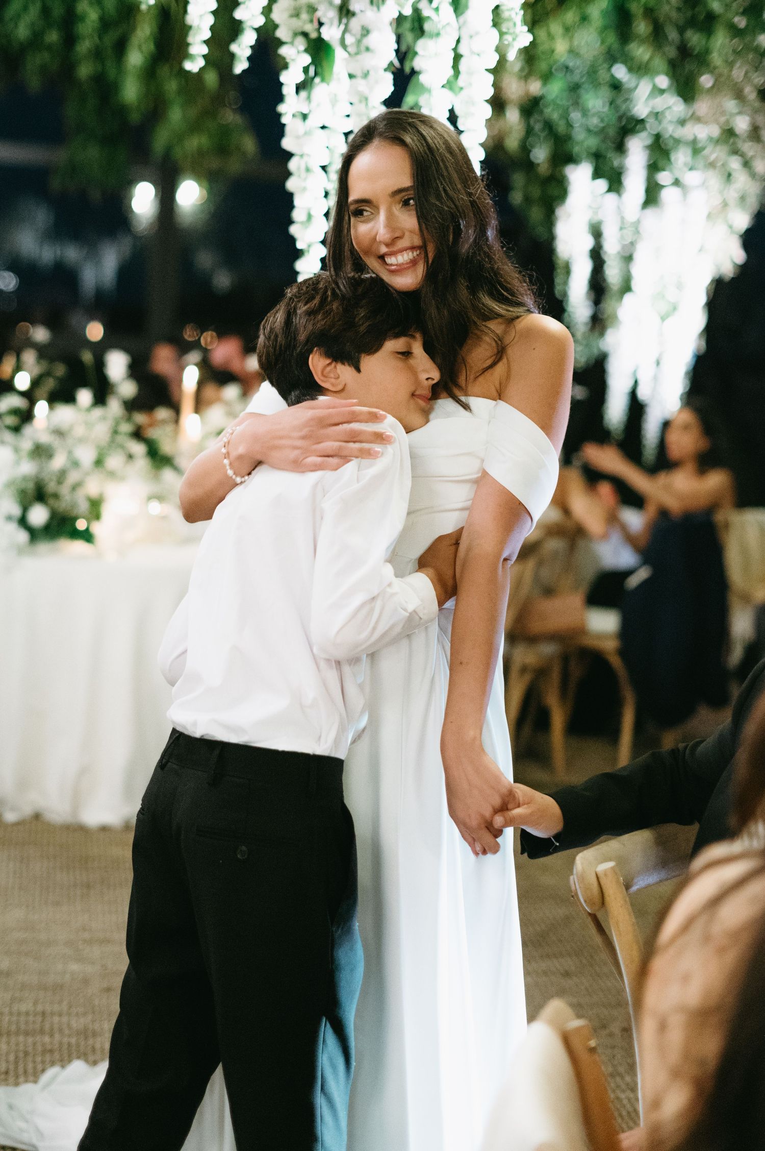 Two people share an emotional embrace during a wedding celebration under hanging floral decorations.