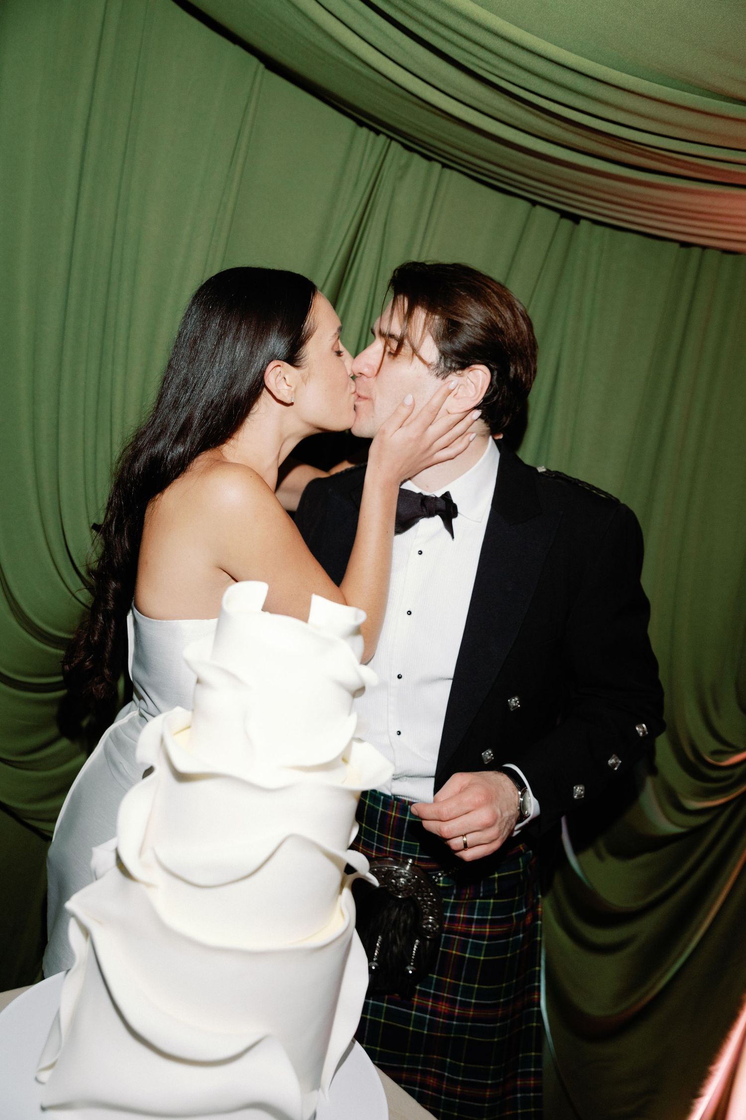 A couple in formal attire sharing a romantic kiss against a green draped backdrop at a special event.