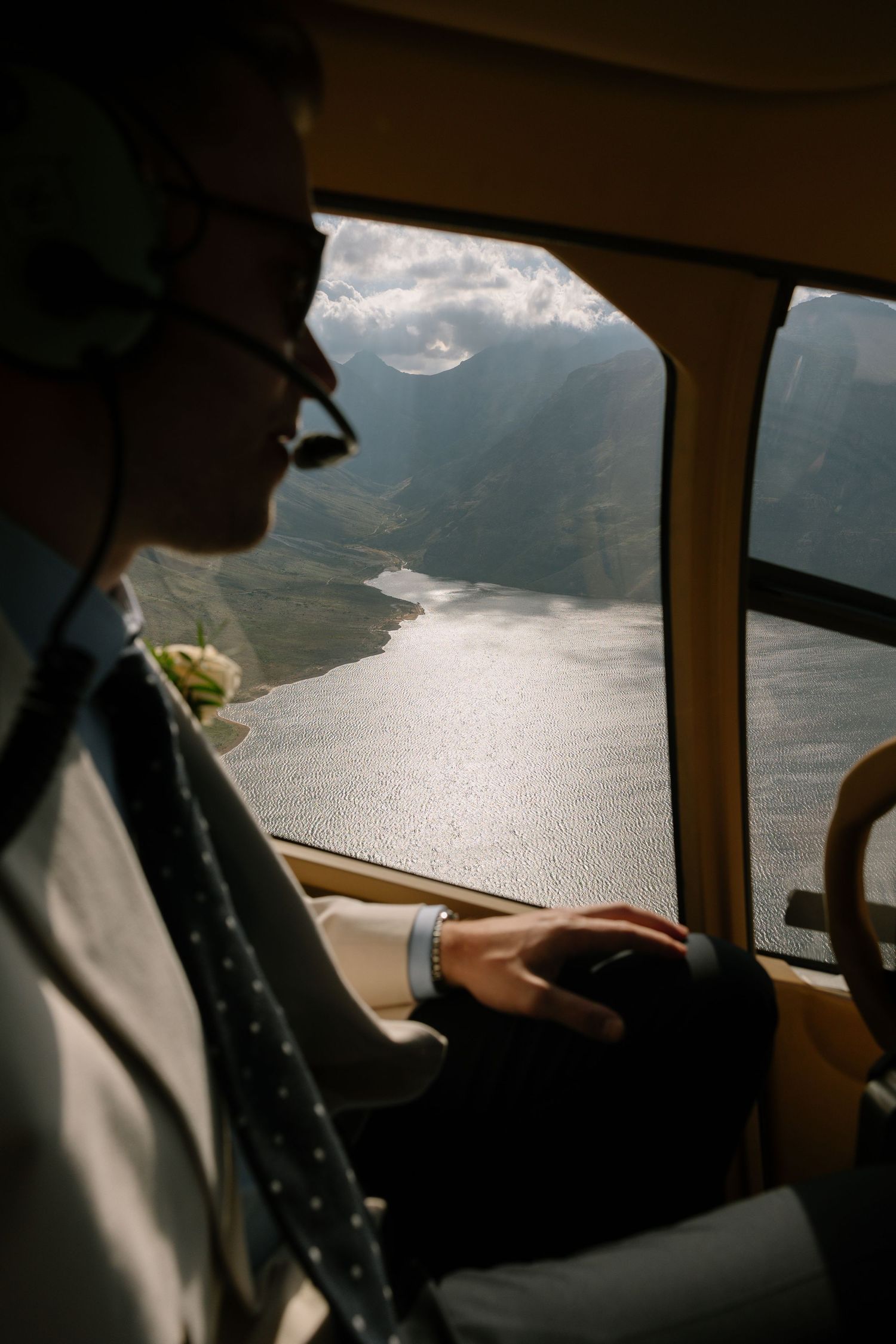 Groom in suit gazes out helicopter window at stunning fjord landscape during scenic wedding day flight.