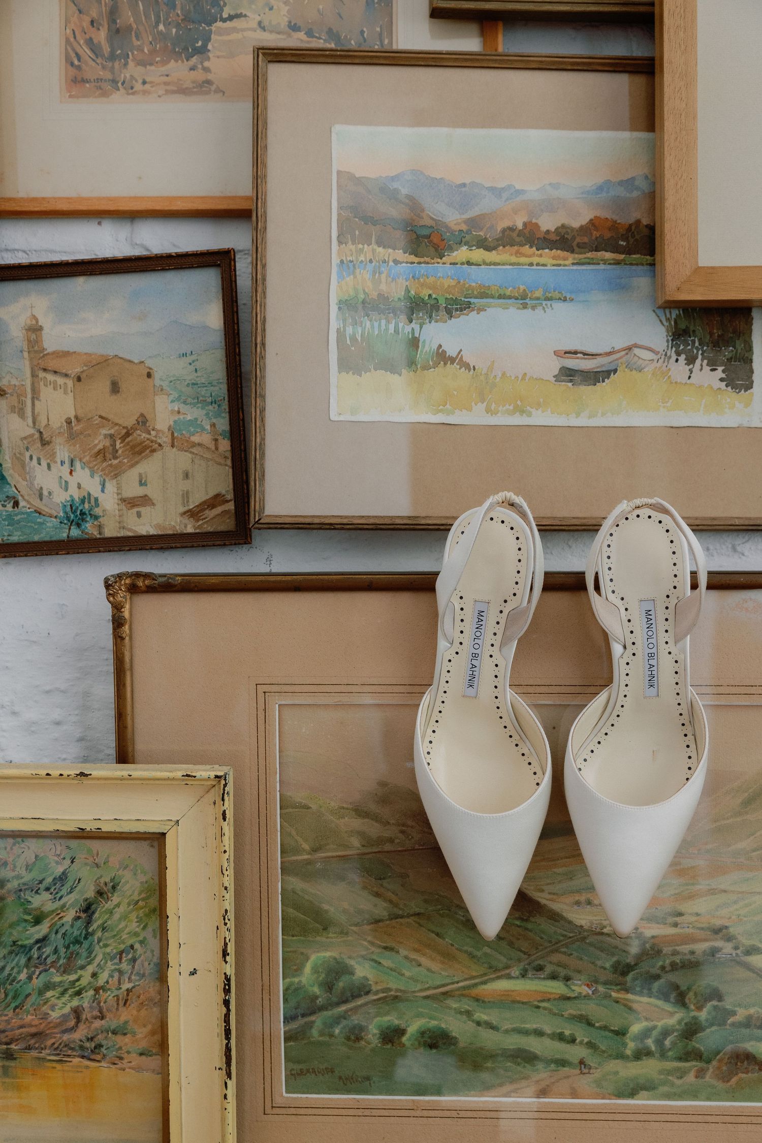 White Manolo Blahnik heels displayed on vintage landscape paintings in ornate frames, perfect bridal shoes for a wedding.
