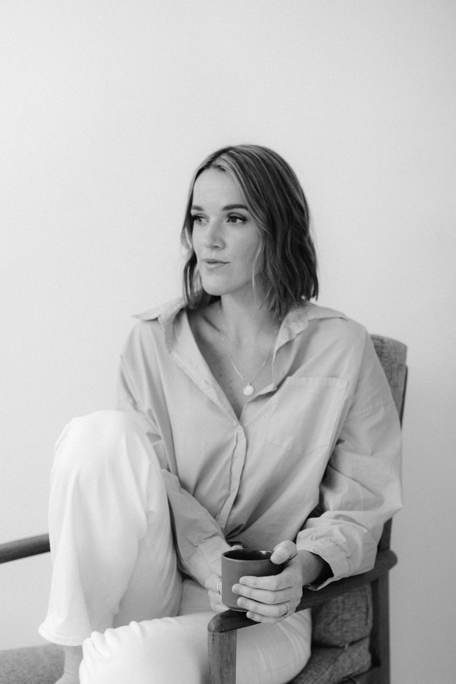 Artistic black and white portrait of a woman in casual attire sitting in a chair holding a cup.