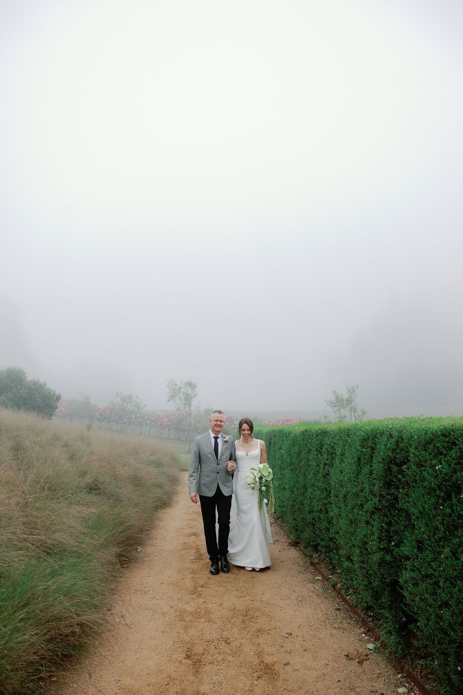 Bride and groom walk a misty fog-covered path lined with hedges at an outdoor vineyard wedding venue.